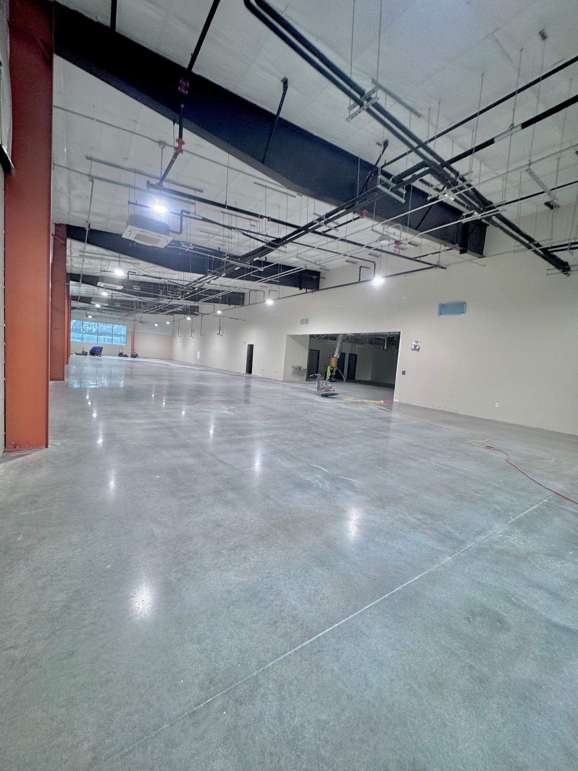 Empty indoor industrial space with polished concrete floor, exposed ceiling pipes, and white walls