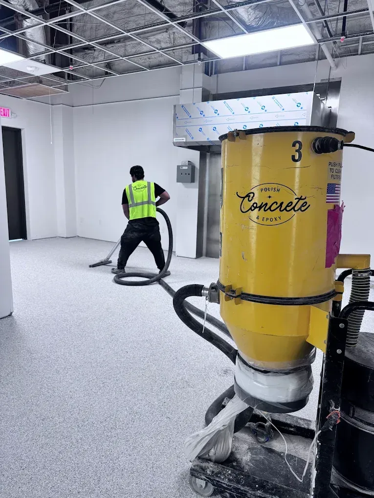 Worker in a neon vest operating a yellow concrete polishing machine in a white indoor space