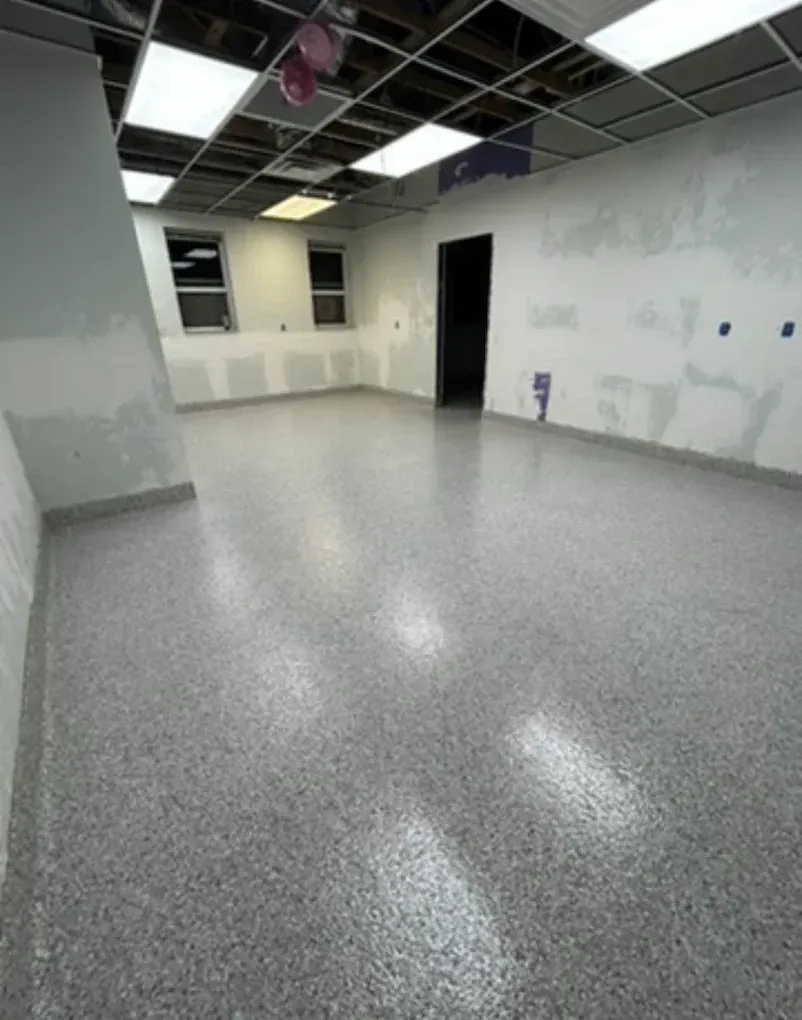 Empty room with speckled gray floor, white walls, and exposed ceiling panels under fluorescent lights