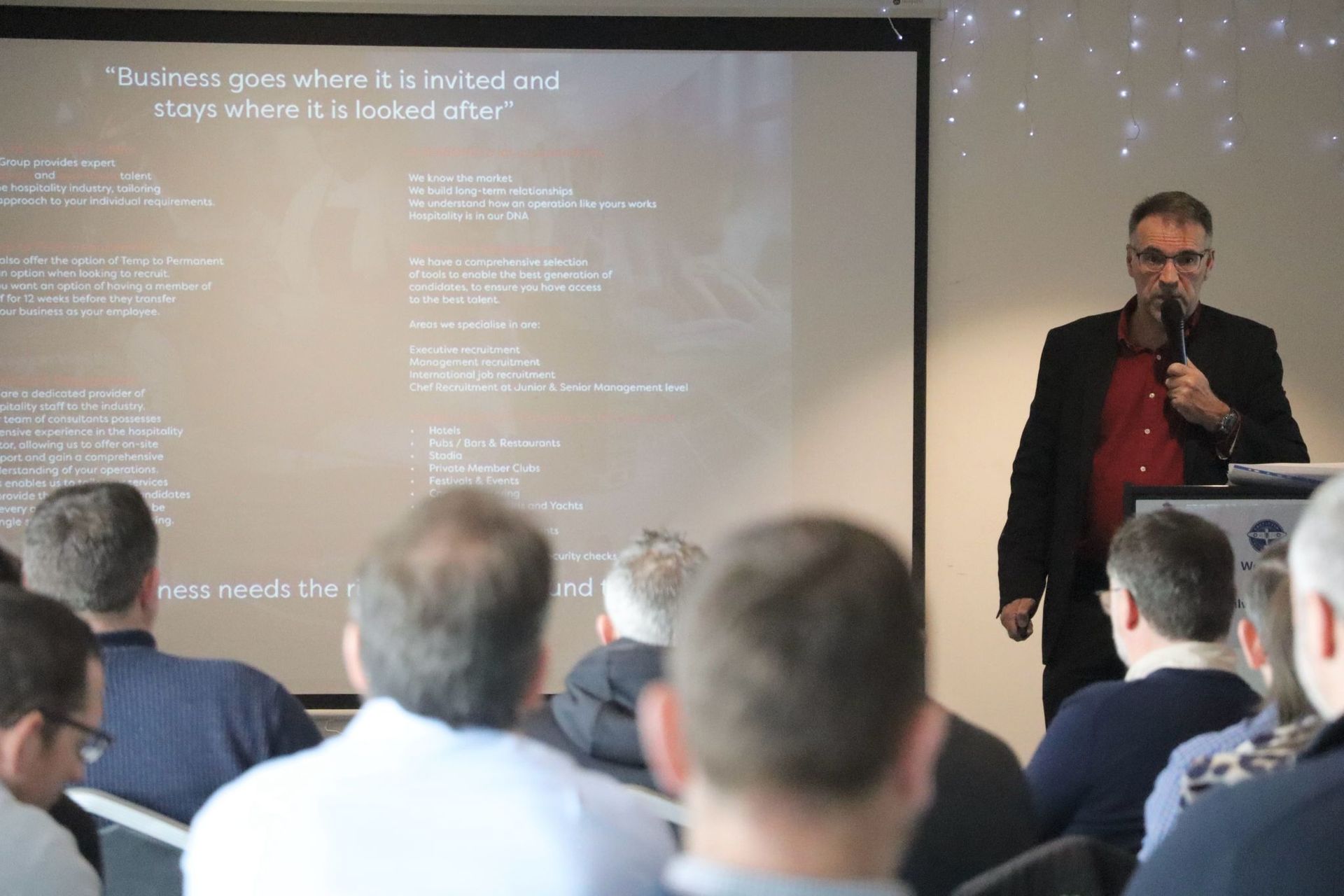 Man speaking at a presentation, screen behind him with text, audience seated.