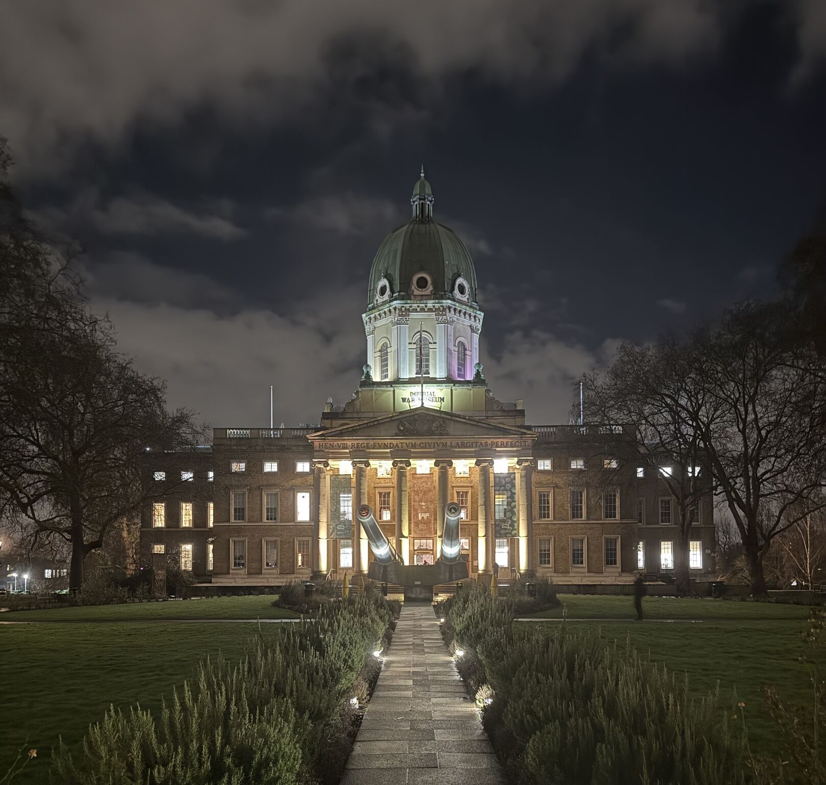 Night view of a lit historic building with a domed tower and central walkway, framed by bare trees