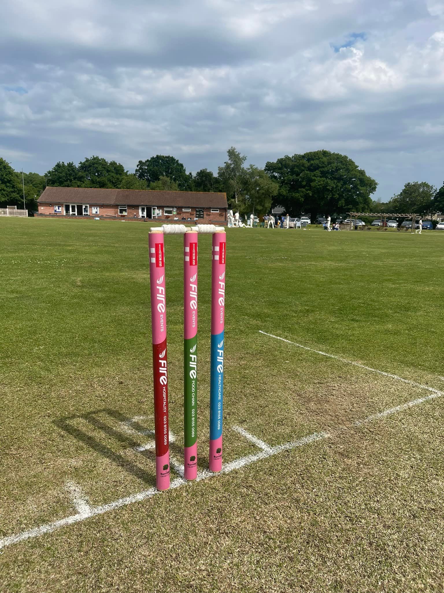 Cricket stumps on a green field, with buildings and trees in the background under a cloudy sky.