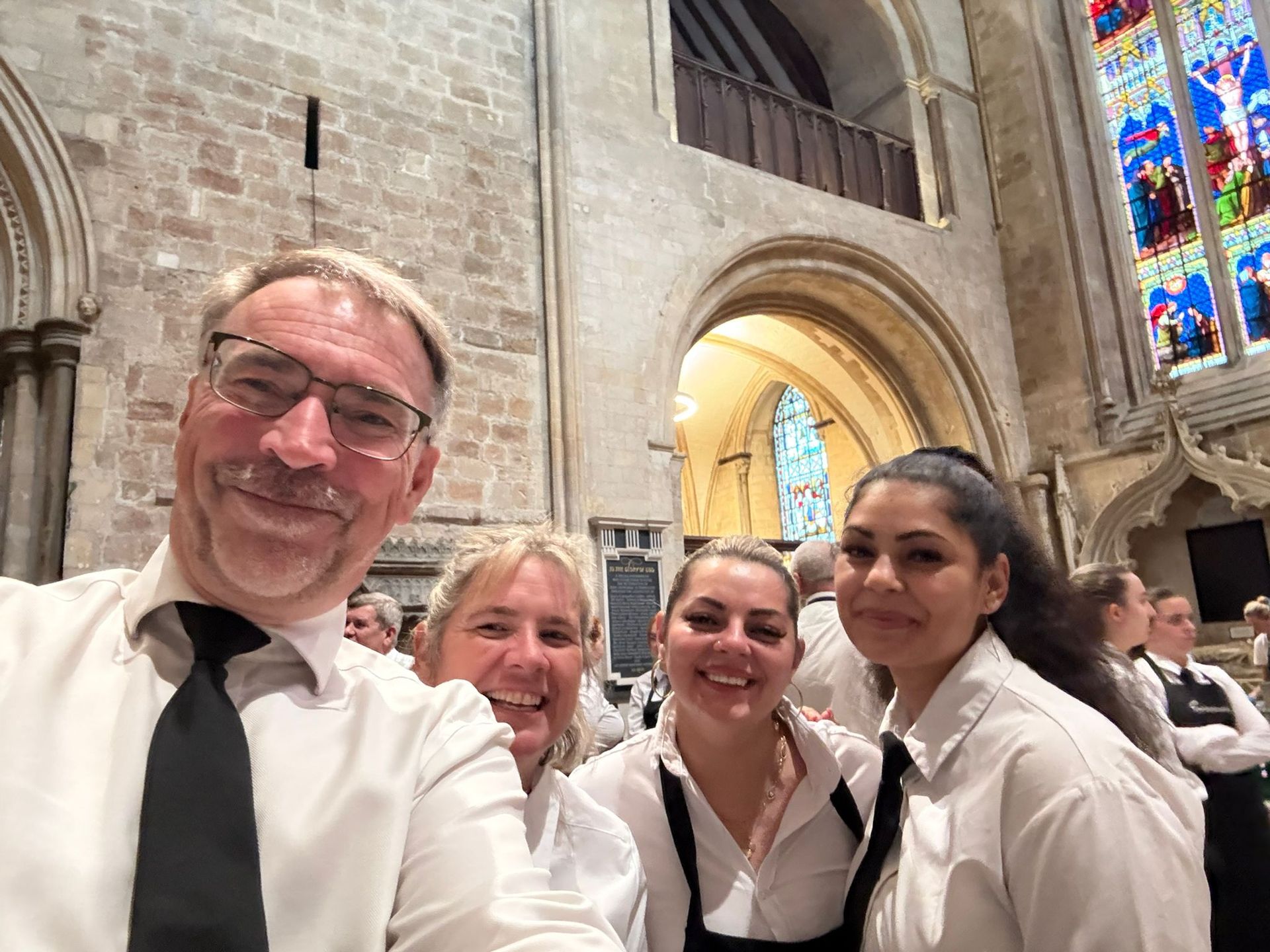 A group of people in white shirts and black ties taking a selfie in a cathedral with stained glass.