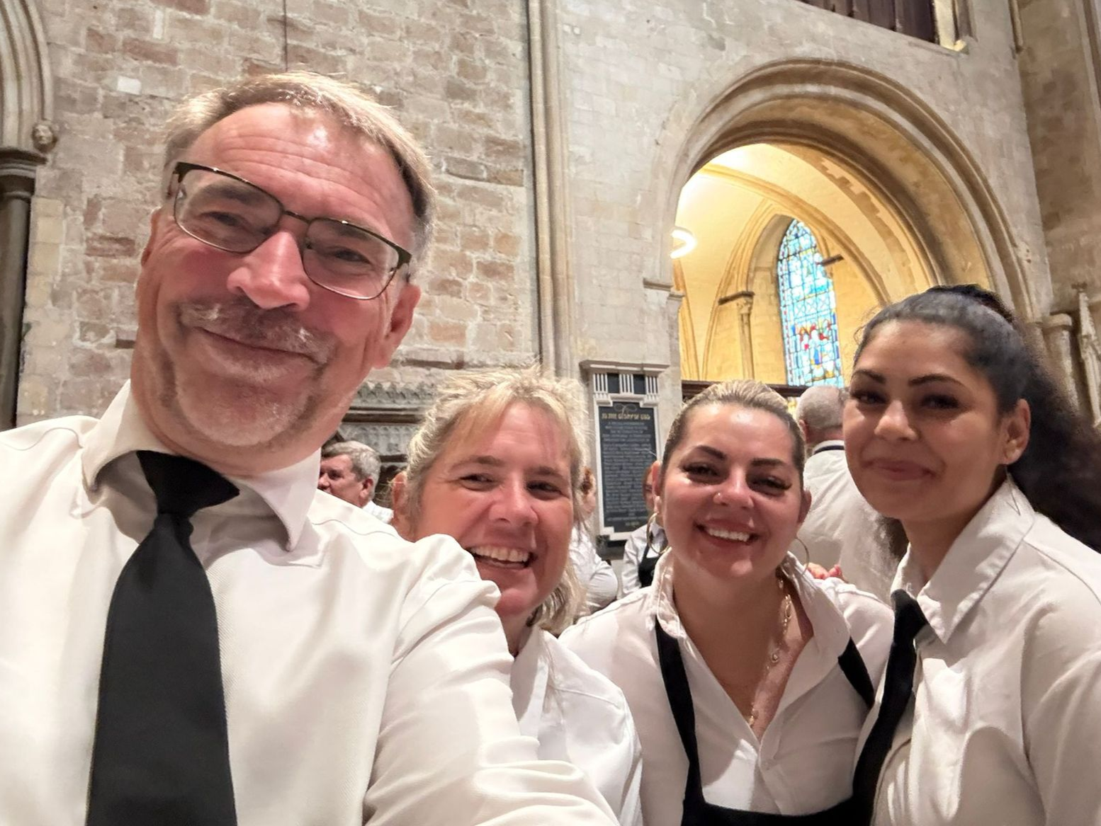 Four smiling people in white shirts and black ties taking a selfie inside a stone building with arched doorways.