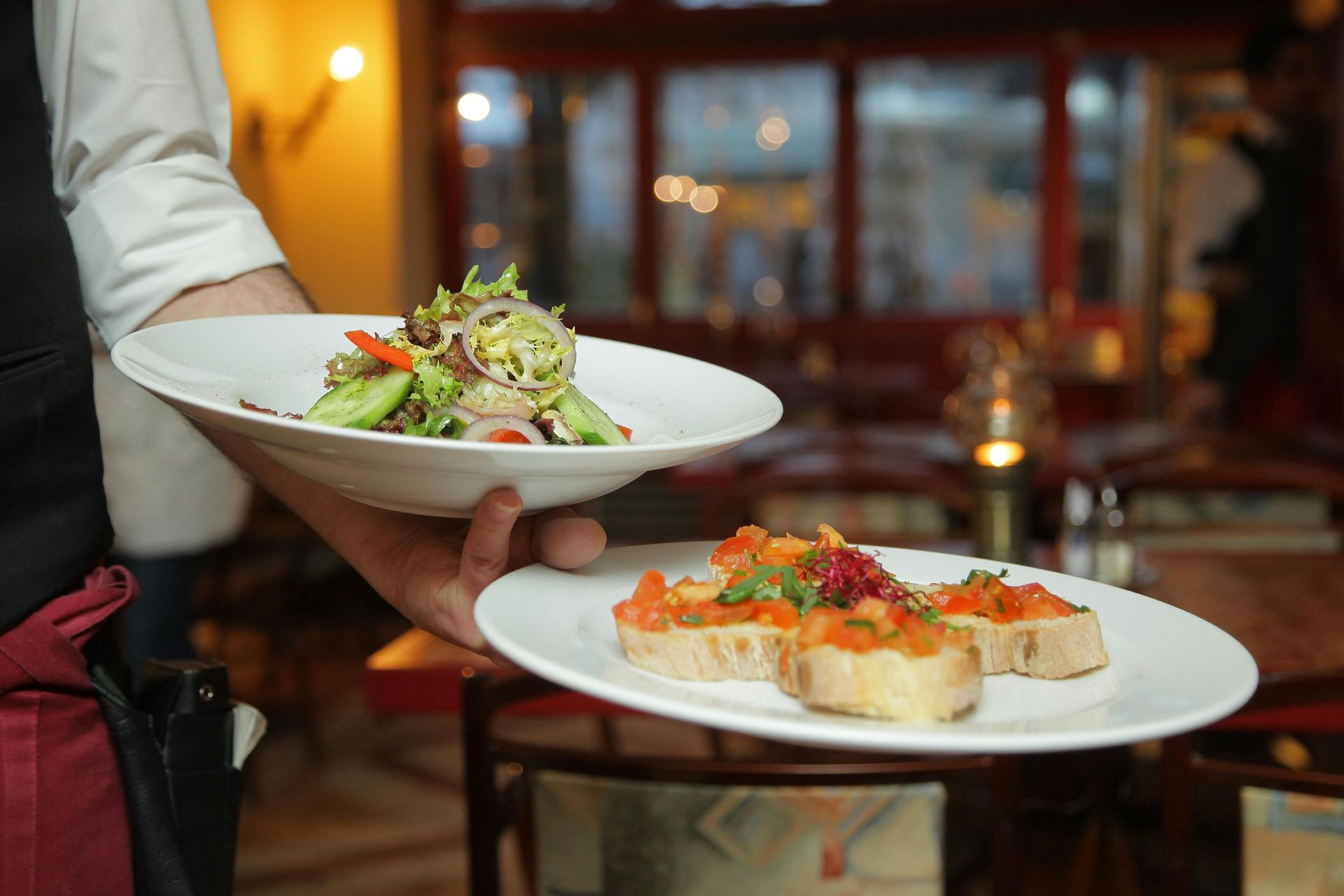 Waiter holding plates of salad and bruschetta in a restaurant setting.