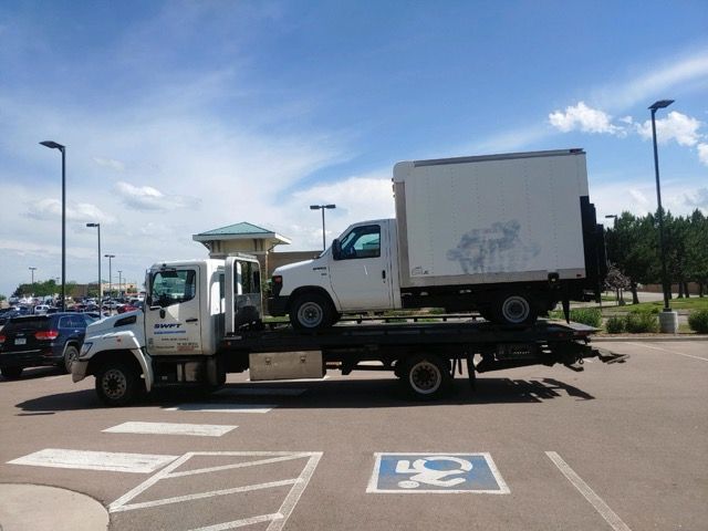 A tow truck is carrying a white truck and a white box