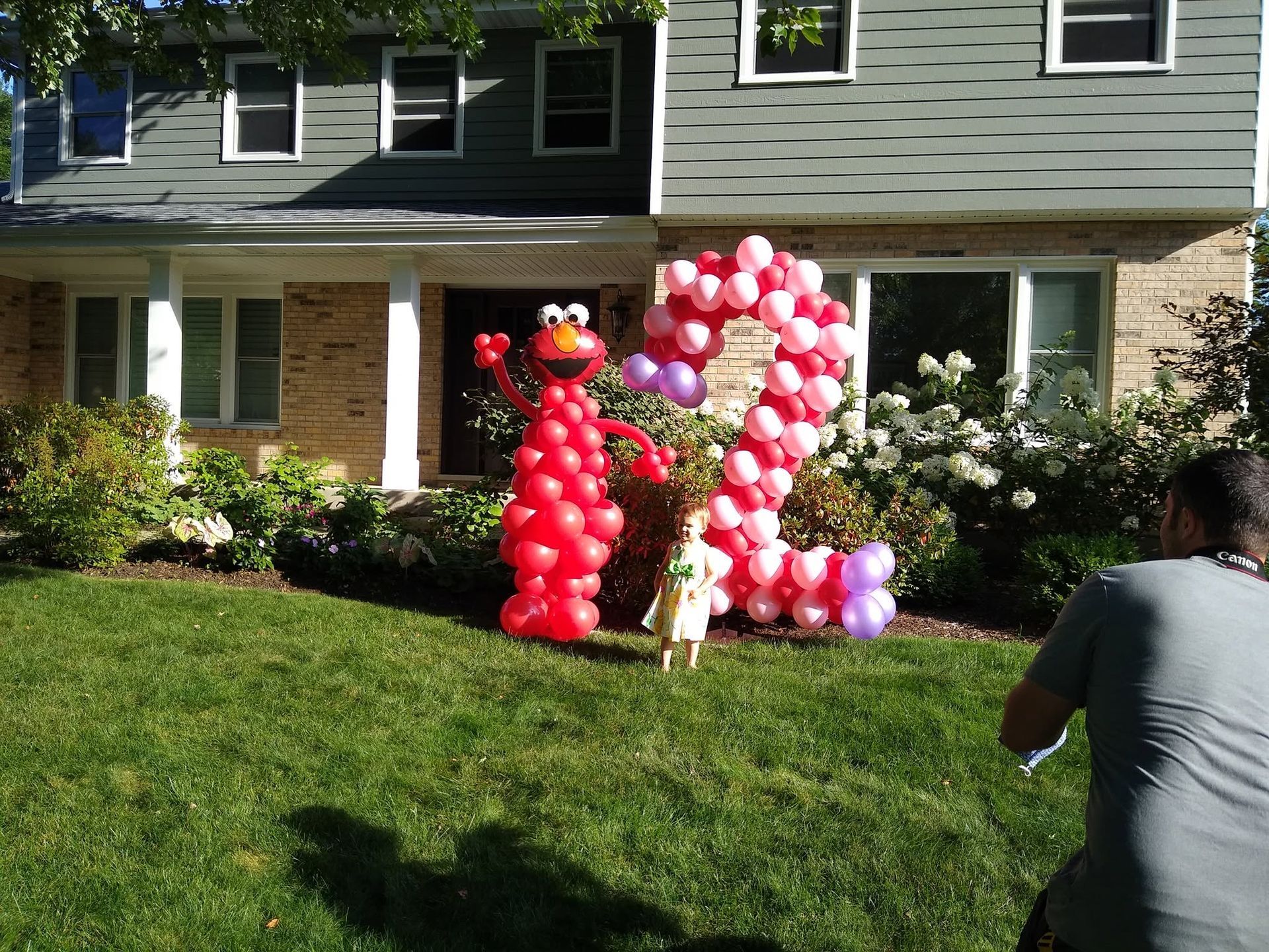 Two balloon sculptures on a lawn in front of a house, with a person filming on the right.