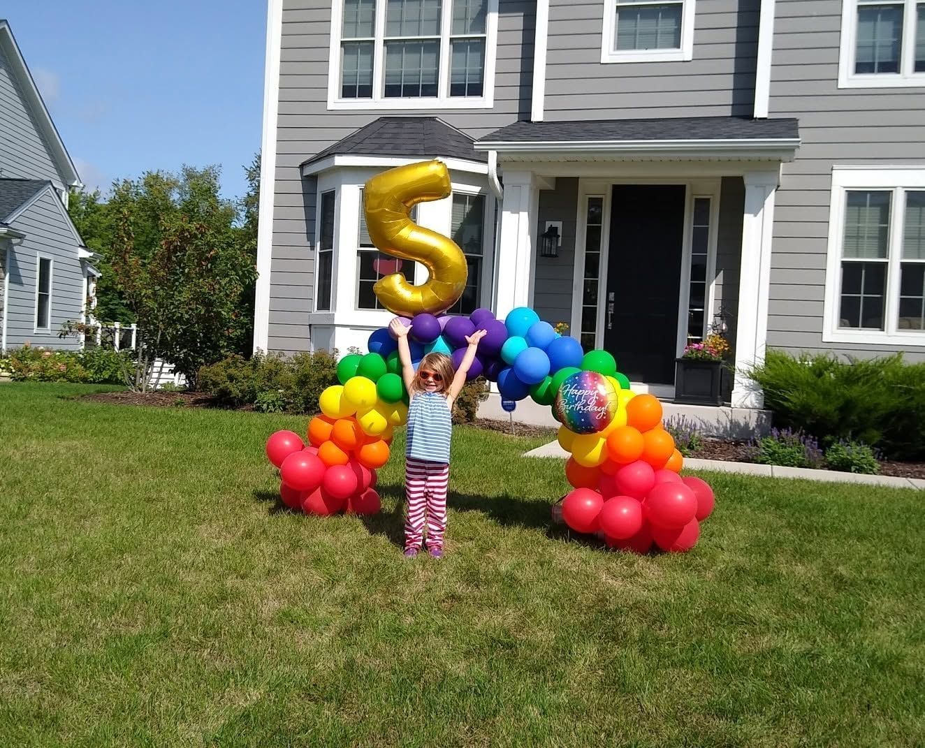Child stands on a lawn with rainbow balloon arch and gold number 5 in front of a house