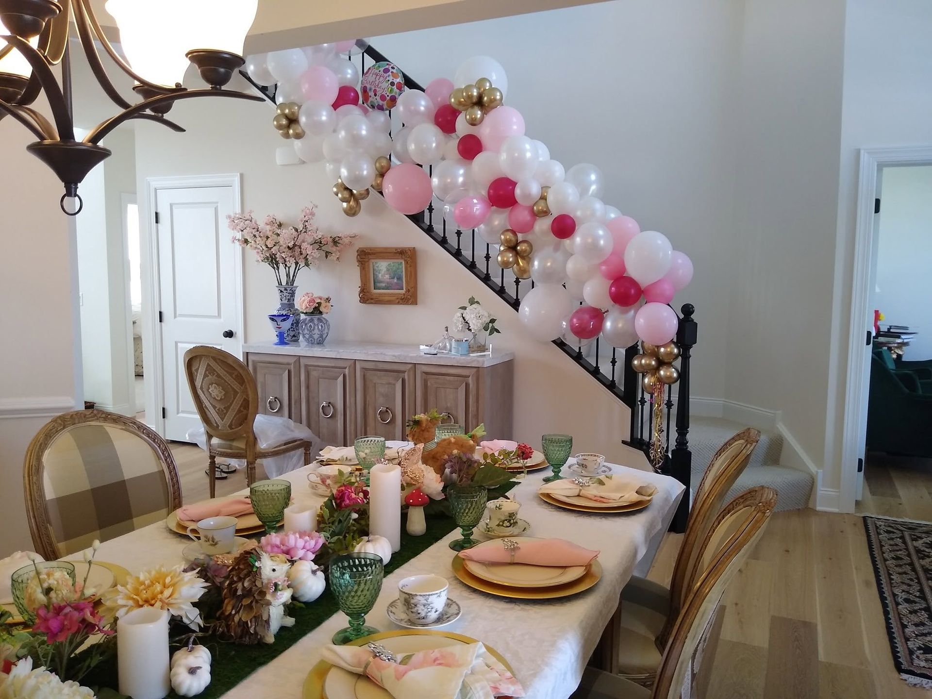 Decorated dining room with a long table, pink and white balloon garland on the staircase, and flowers on the table