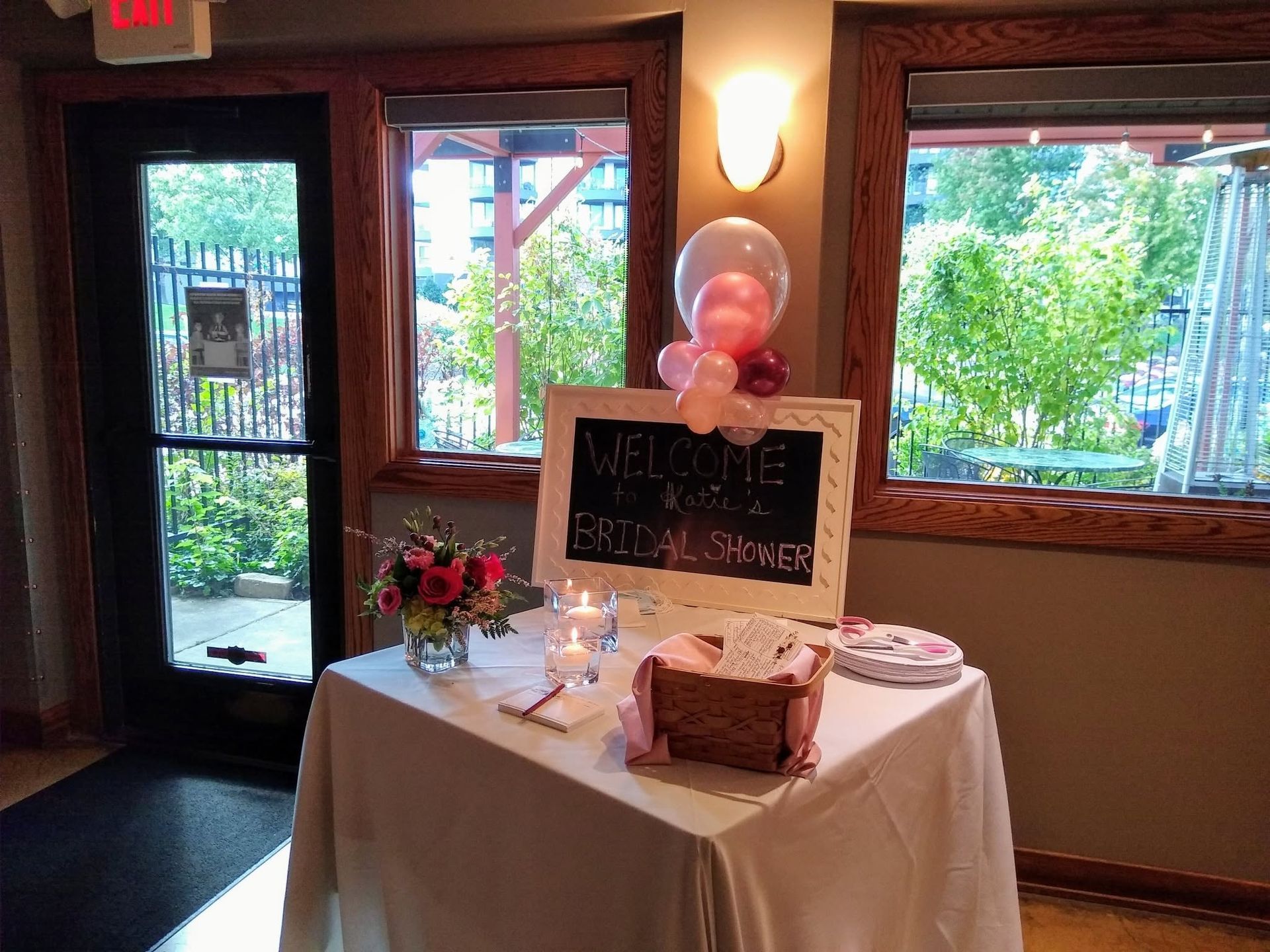 Decorated table with chalkboard sign and balloons in a warmly lit lobby by glass doors