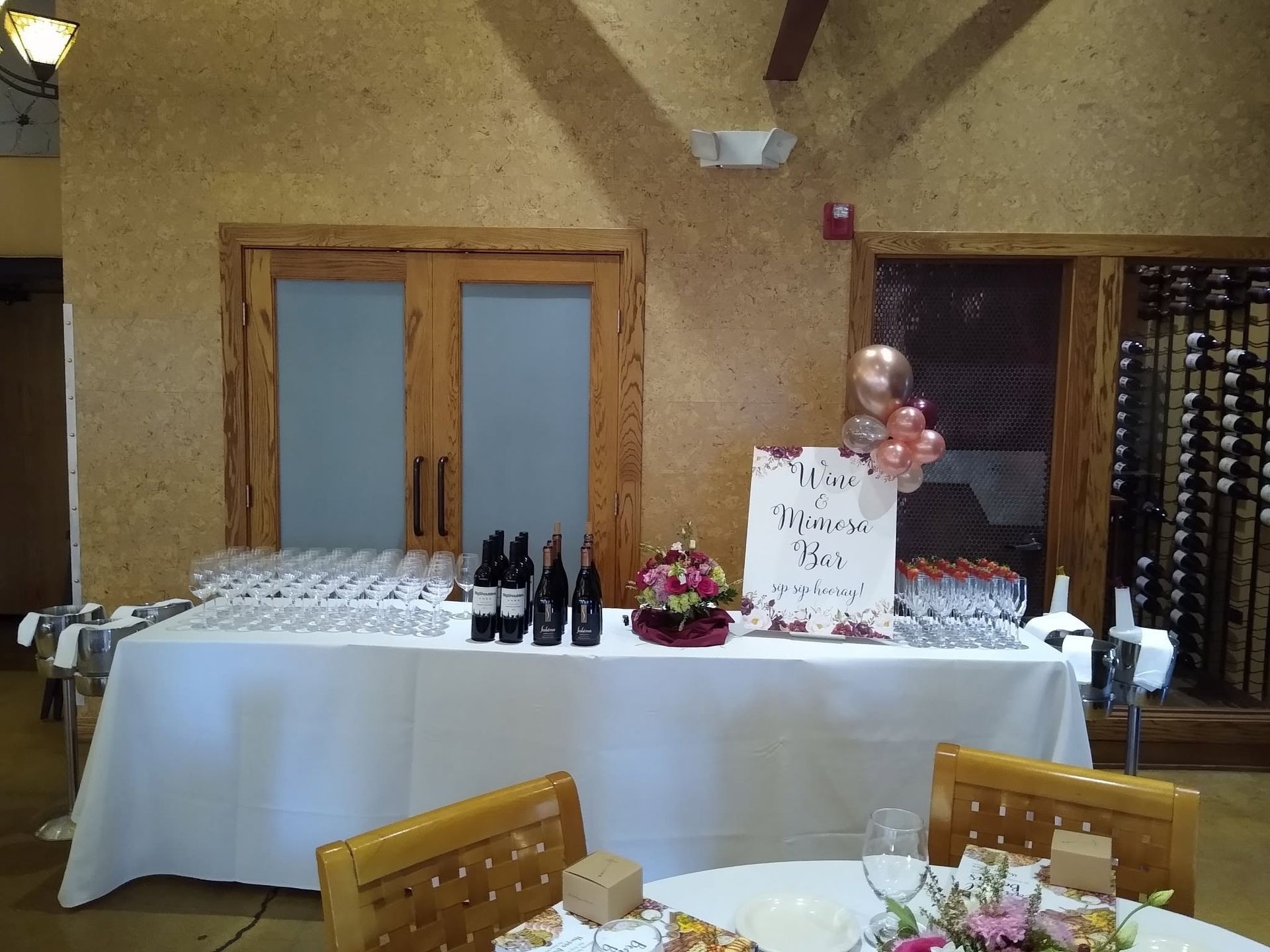 Decorated banquet table with wine bottles, flowers, balloons, and a sign in a hall