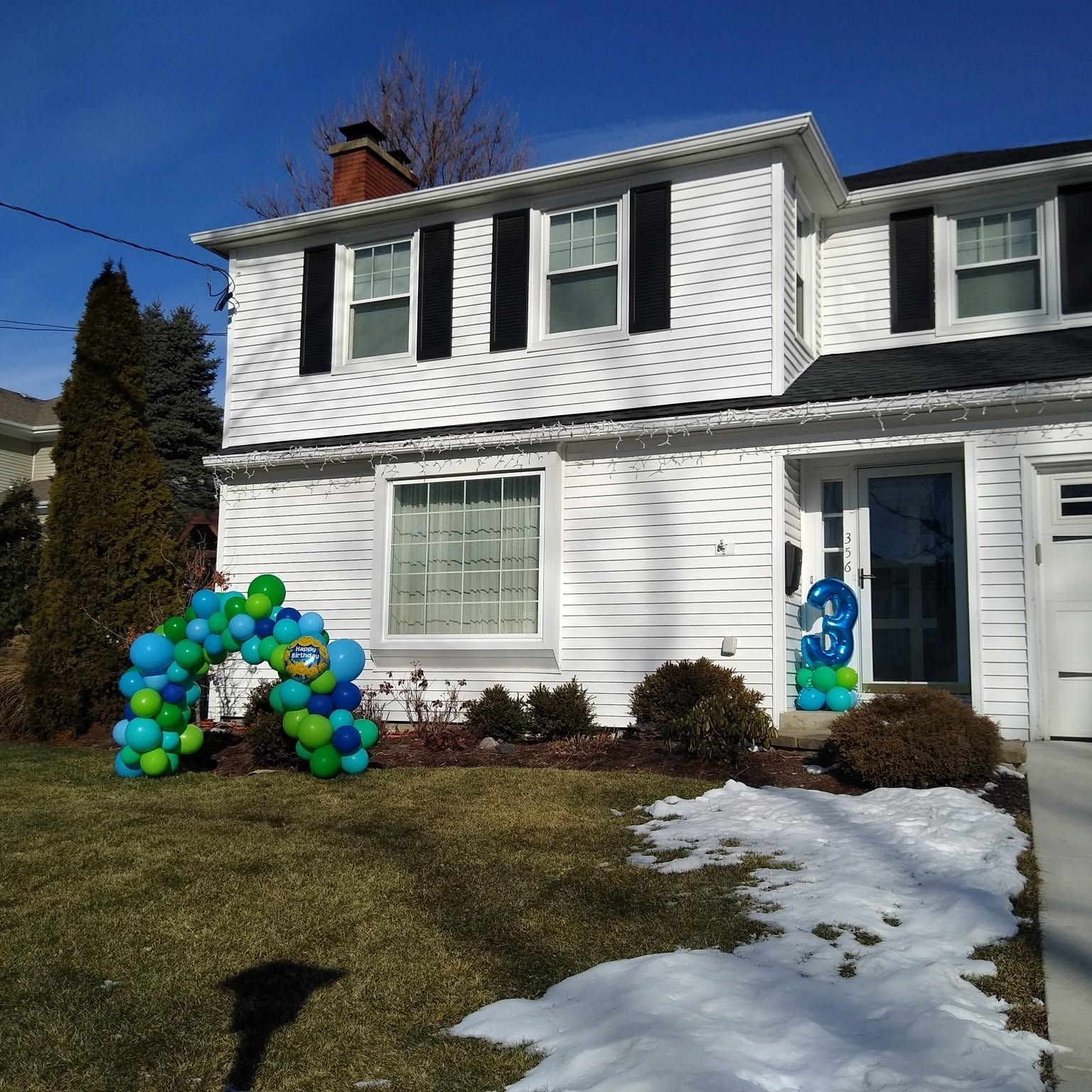 White two-story house decorated with blue and green balloon arch near the front door, with snow on the lawn.