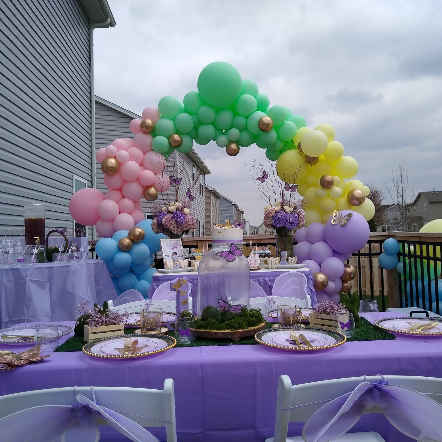 Colorful balloon arch over a lavender outdoor party table with plates and floral centerpieces