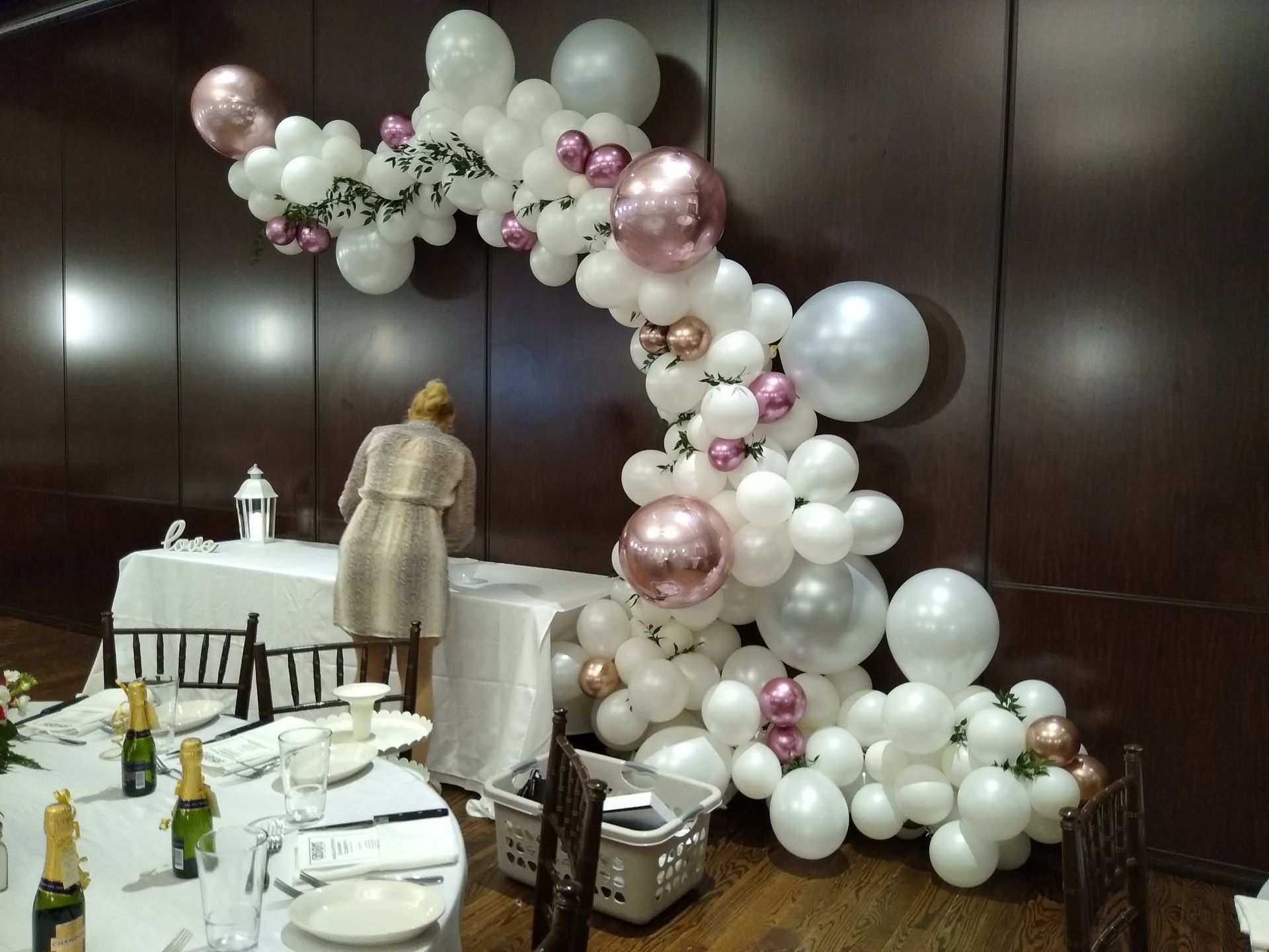 Balloon arch and decorated banquet table in a dark wood room, with a person arranging table settings