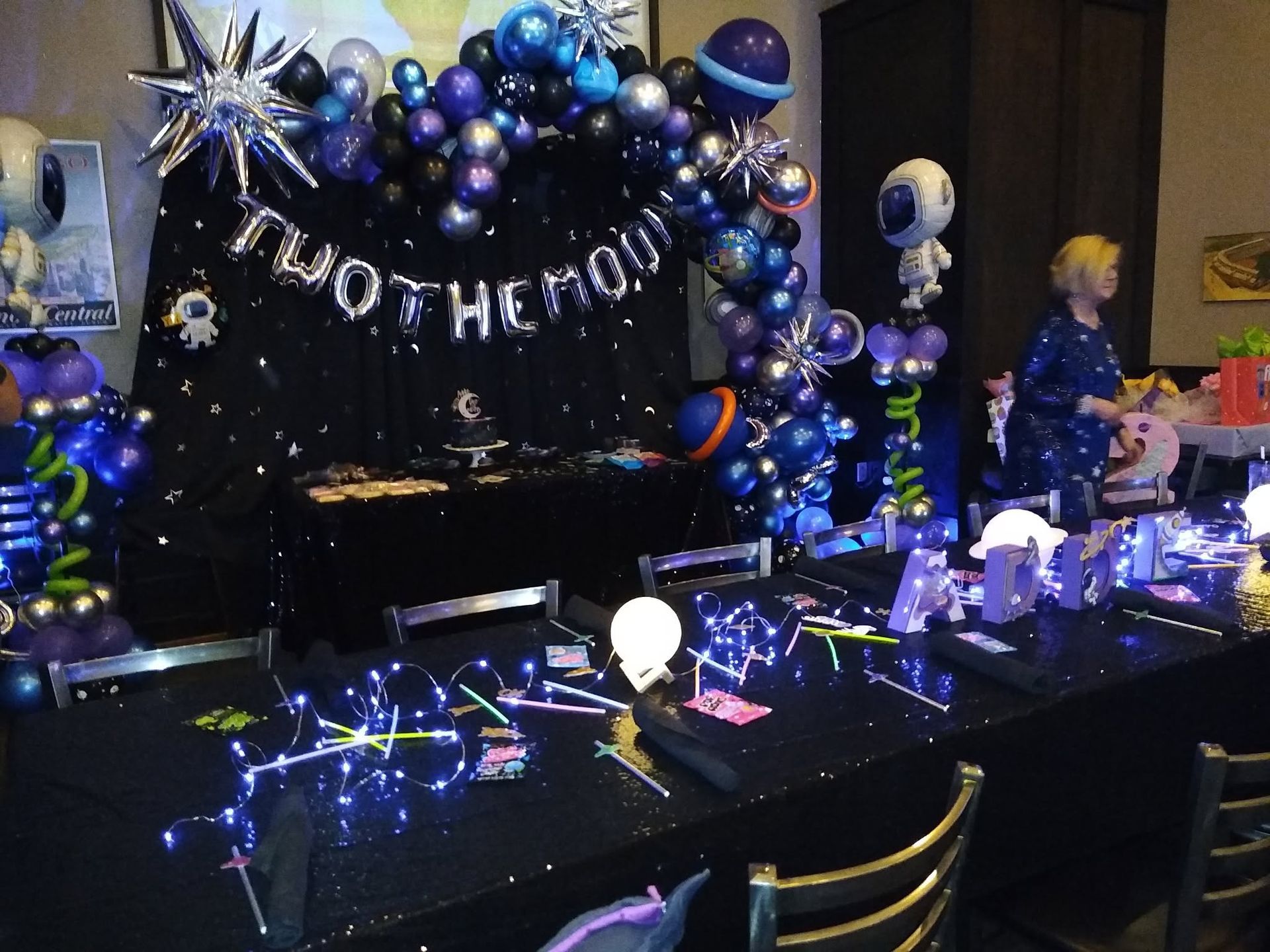 Black and silver birthday party table with balloon arch, gift bags, lights, and a “Happy Birthday” banner