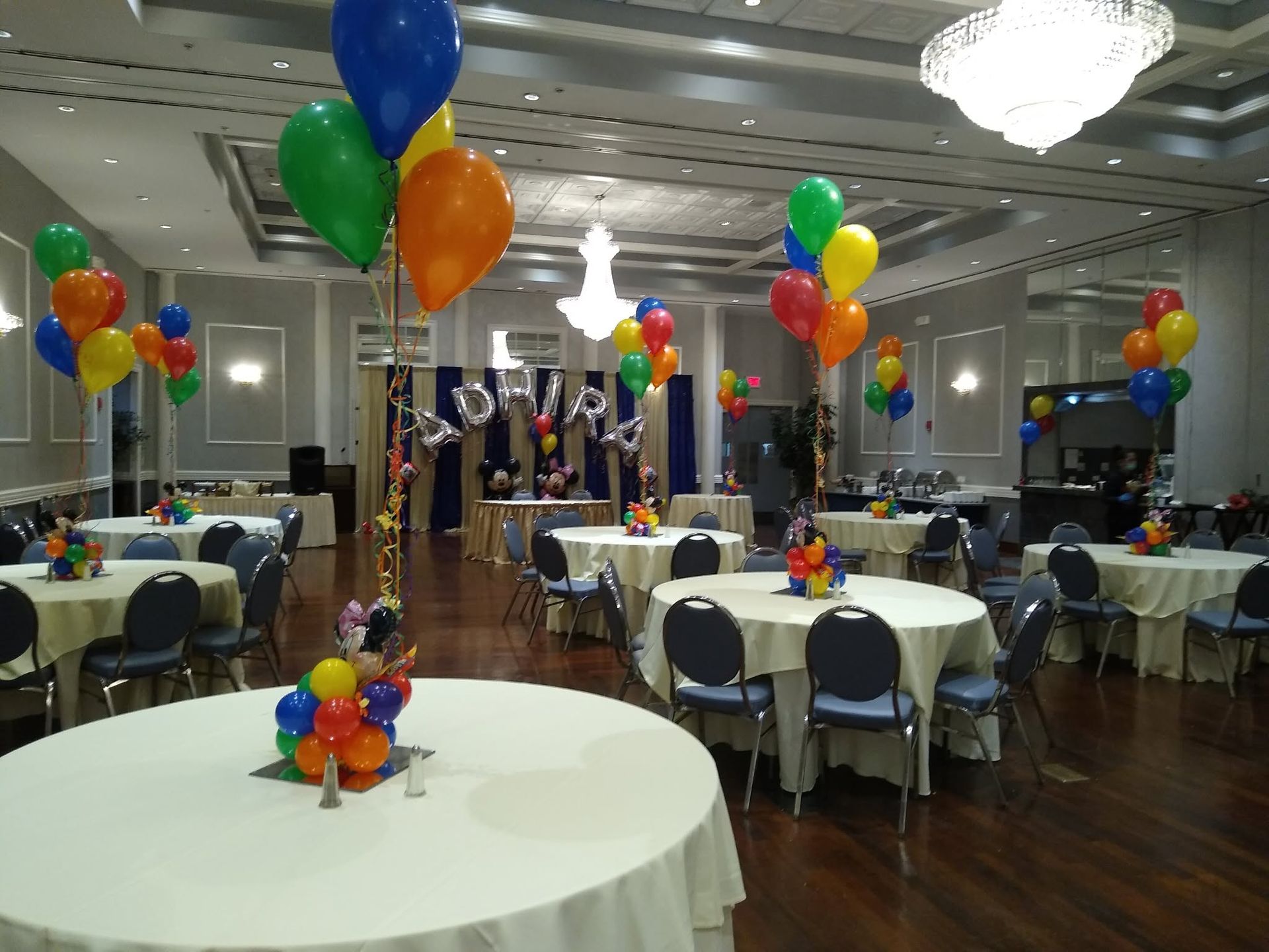 Balloon-decorated banquet hall with round tables, chairs, and chandeliers, set for a celebration