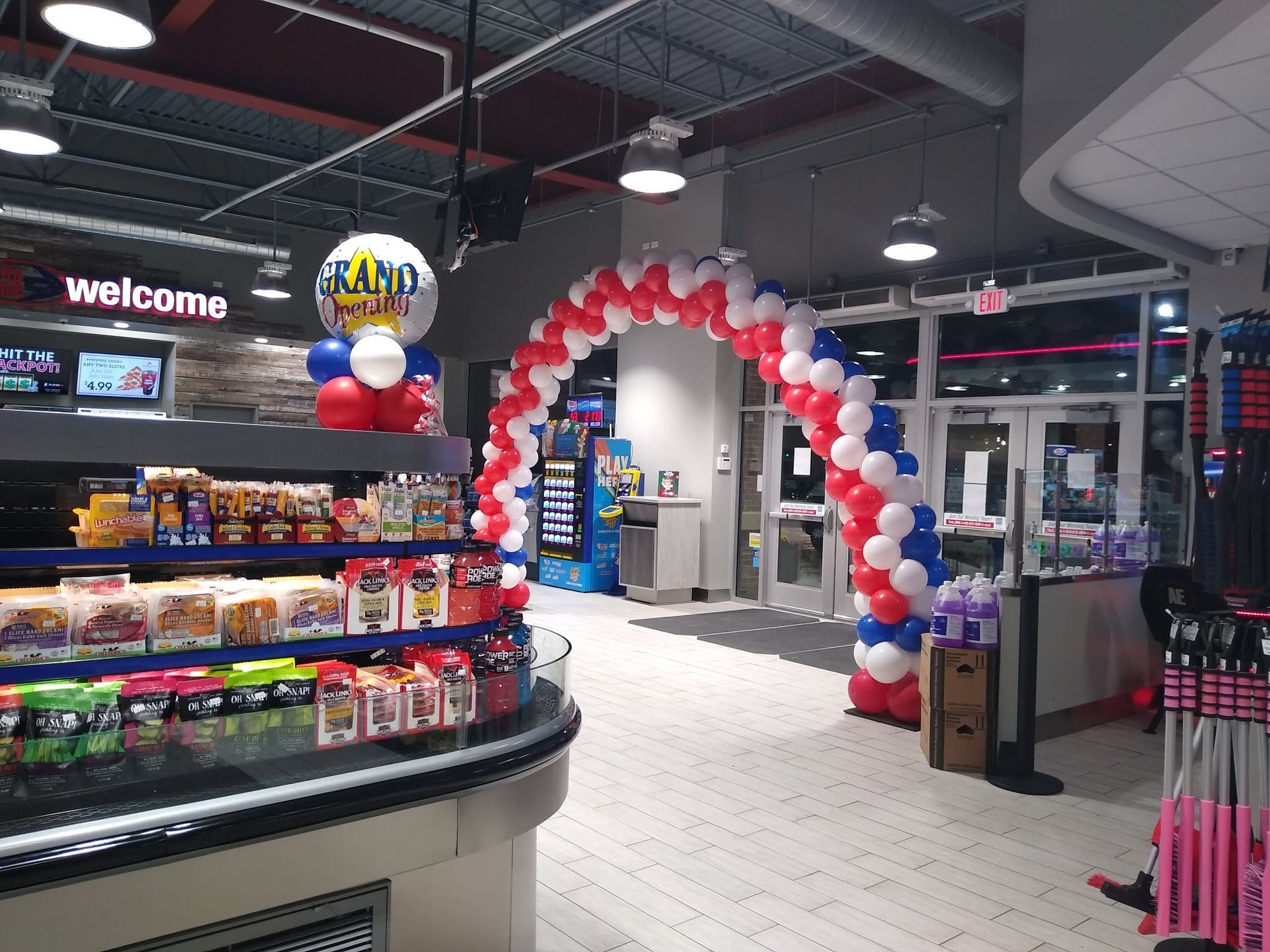 Store entrance with a red, white, and blue balloon arch and a “welcome” sign by candy shelves
