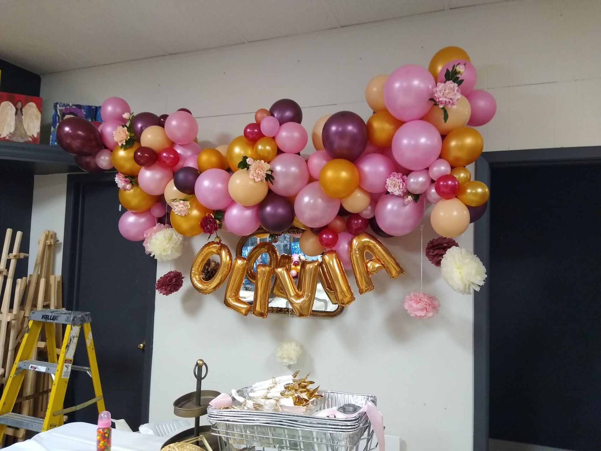Pink, gold, and white balloon backdrop with “GRACIA” letters and a decorated table beneath.