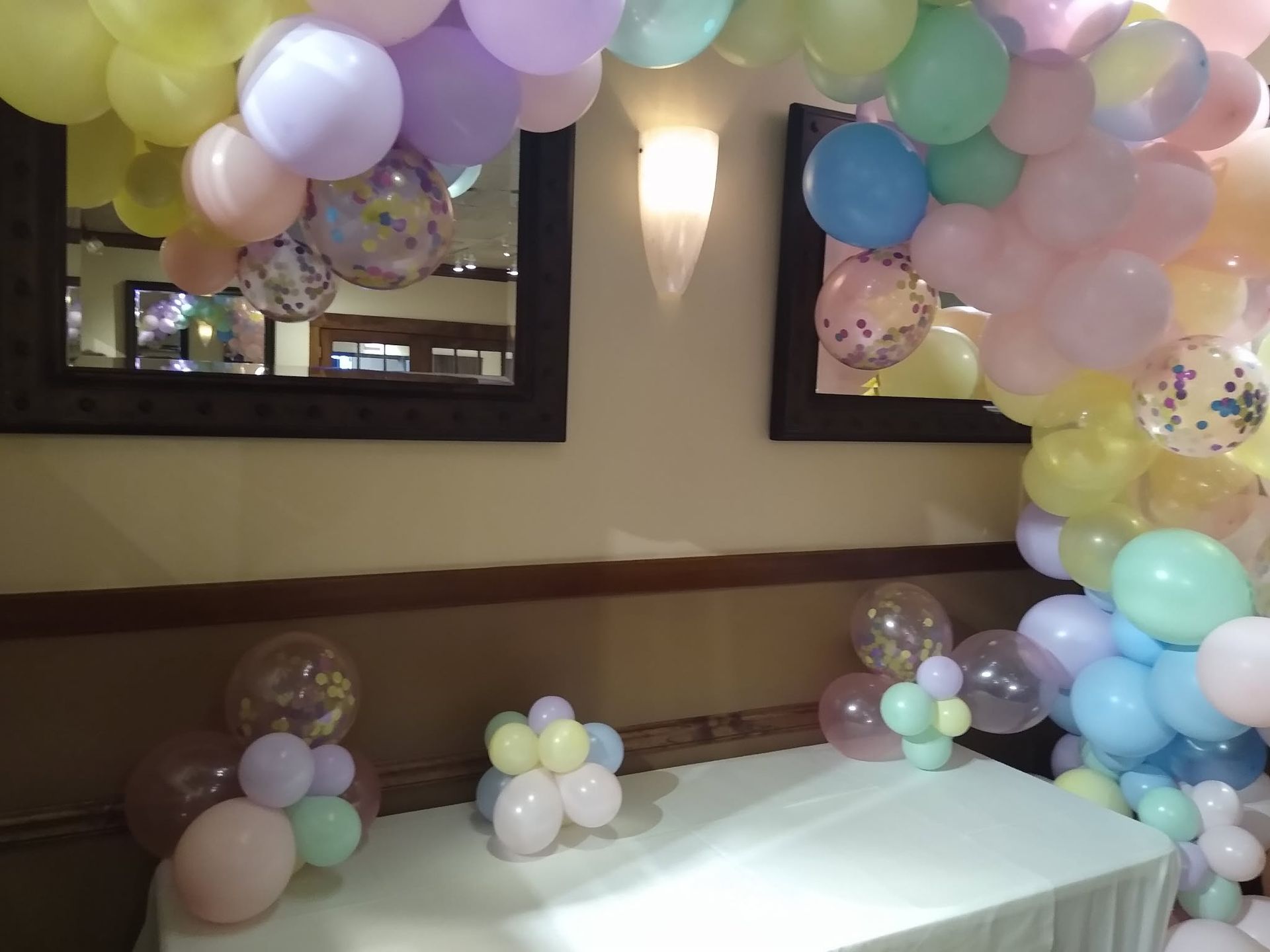 Pastel balloon arch and clusters decorating a banquet room with a white table and wall sconces