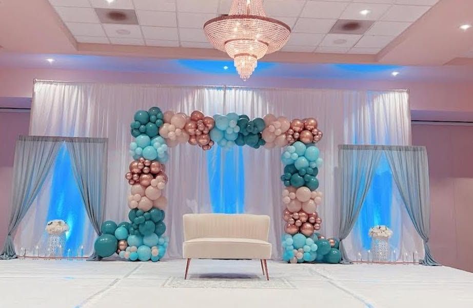 Elegant event stage with teal and blush balloon arch, white loveseat, and draped blue-lit backdrops