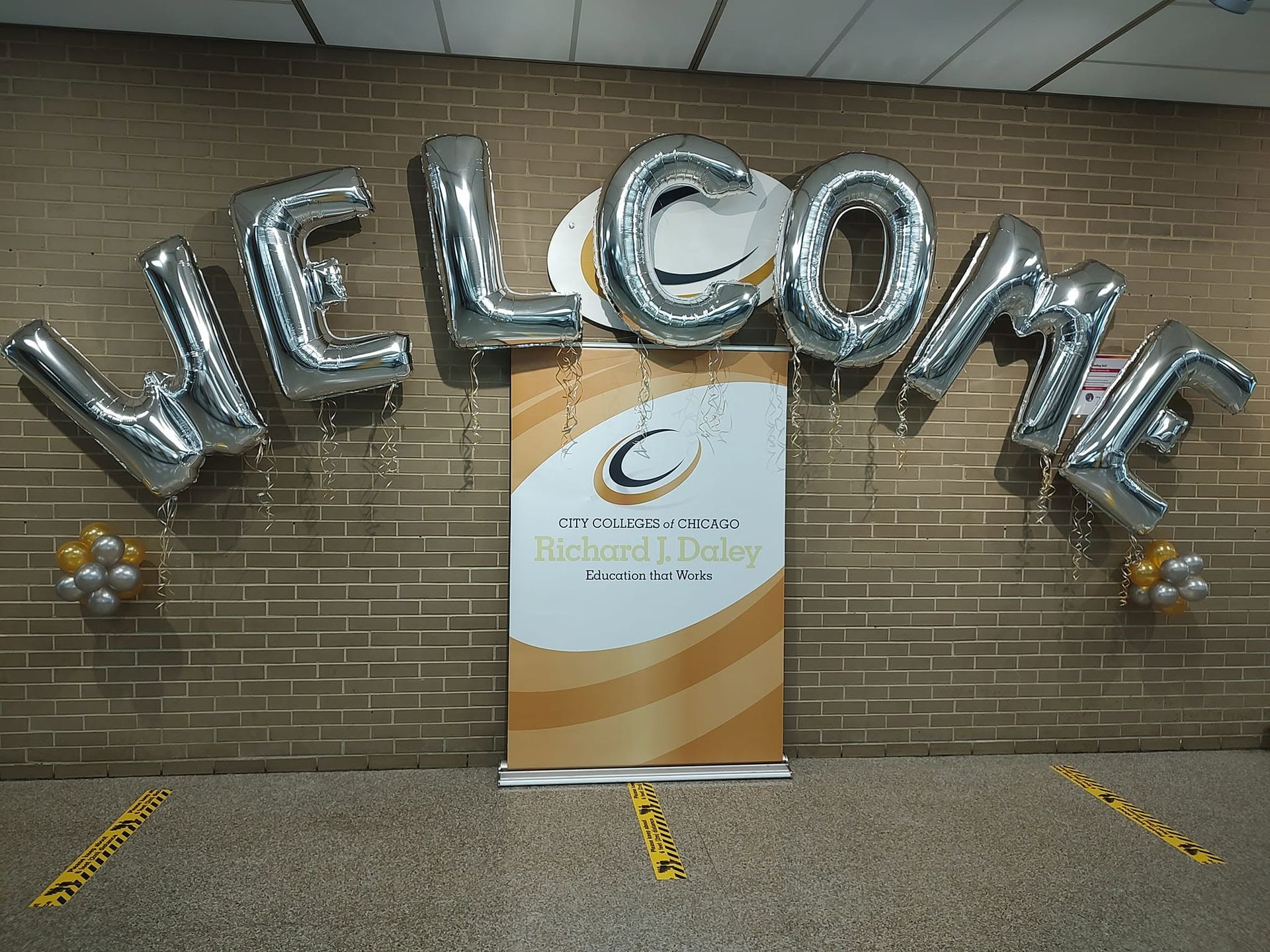 Silver balloon letters spelling “WELCOME” above a sign on a brick wall indoors.