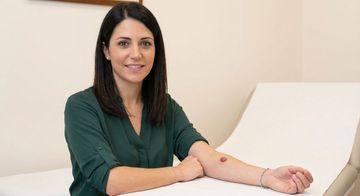 Donna seduta su una sedia bianca, sorridente, con indosso una camicetta verde e un segno rosso sull'avambraccio.