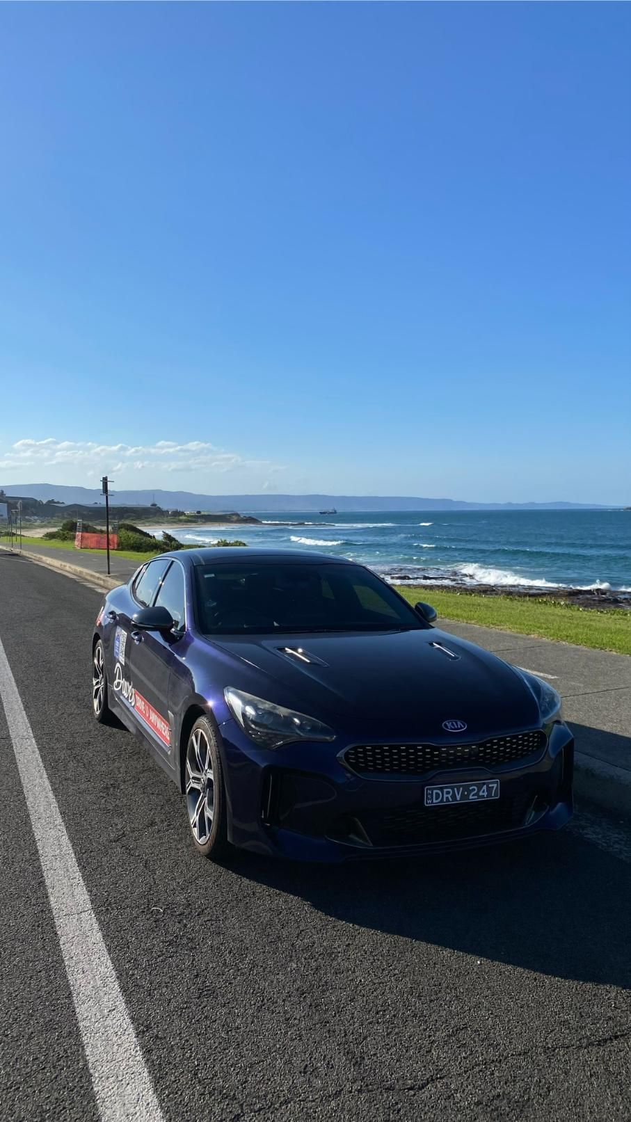 Blue KIA parked on a coastal road with the ocean in the background under a clear blue sky — Dave's Drive U Anywhere 24/7 Mini Van Hire In Albion Park, NSW