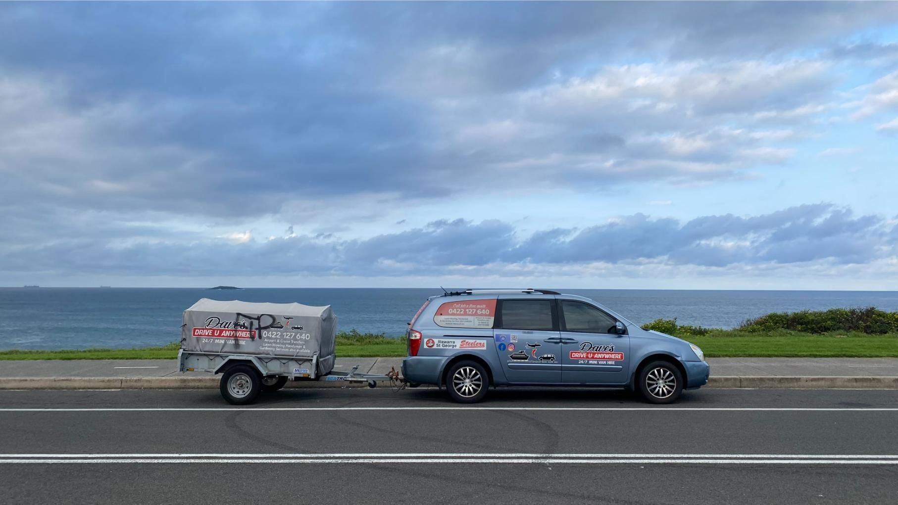 Blue car towing a trailer on a road with ocean and cloudy sky in background. — Dave's Drive U Anywhere 24/7 Mini Van Hire In Bulli, NSW