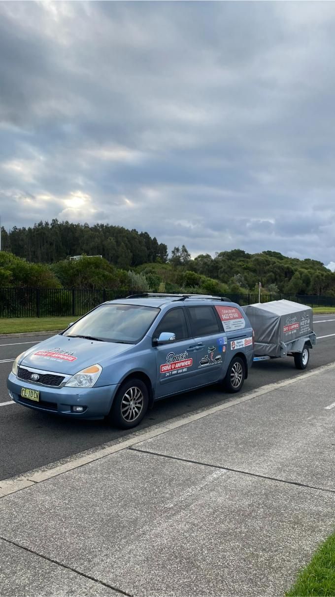 Blue minivan towing a small trailer, on a road under a cloudy sky, trees in background — Dave's Drive U Anywhere 24/7 Mini Van Hire In Kiama, NSW