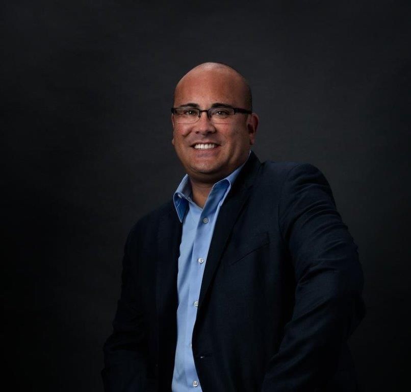 A man wearing glasses and a dark blazer over a light blue button-down shirt, smiling against a dark background.