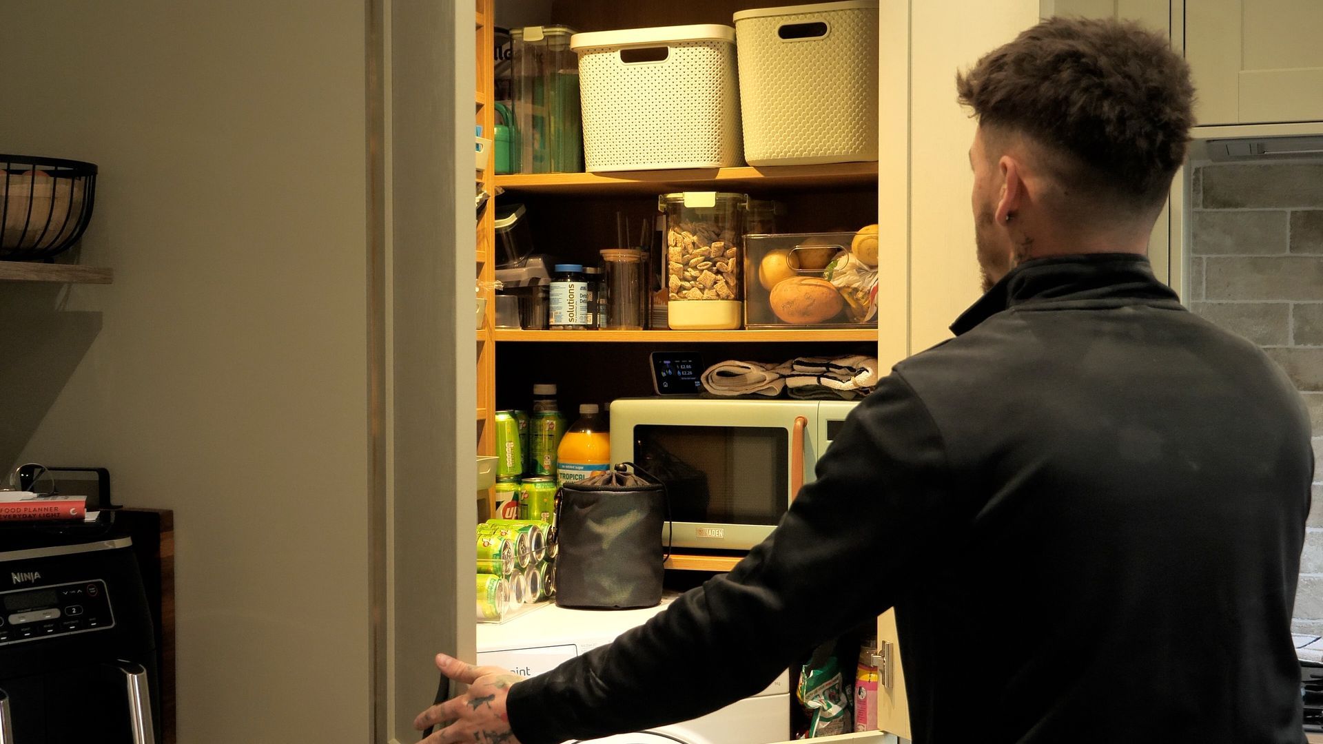 Man opening a pantry door filled with food and containers. Interior kitchen setting.