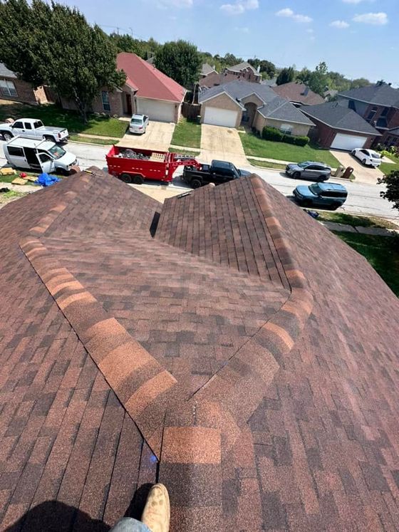 Roof shingles and ridge line viewed from a rooftop, overlooking a suburban street and houses below