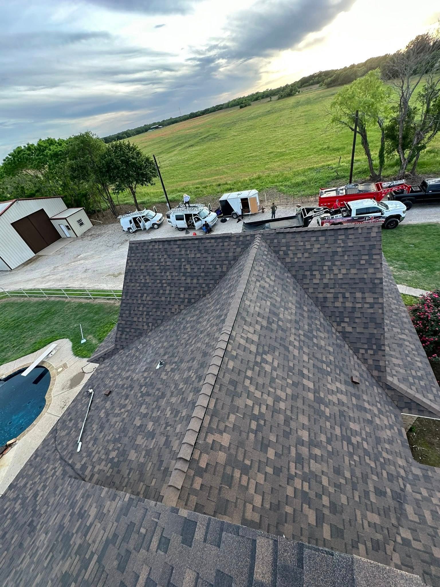 Roofline overlooking a rural yard with parked vehicles, trees, and rolling green hills under a cloudy sky