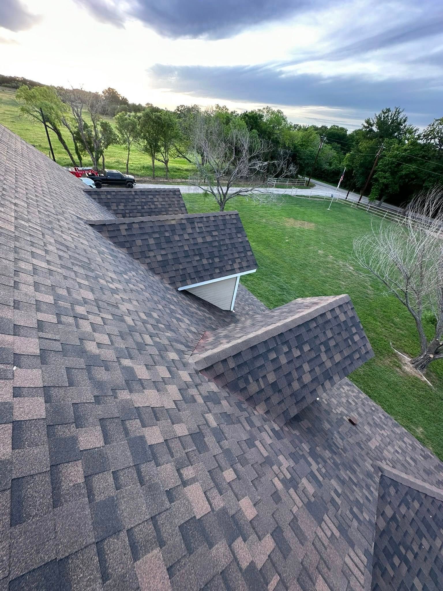 Roof shingles in foreground overlooking a grassy yard, trees, and cloudy sky at dusk