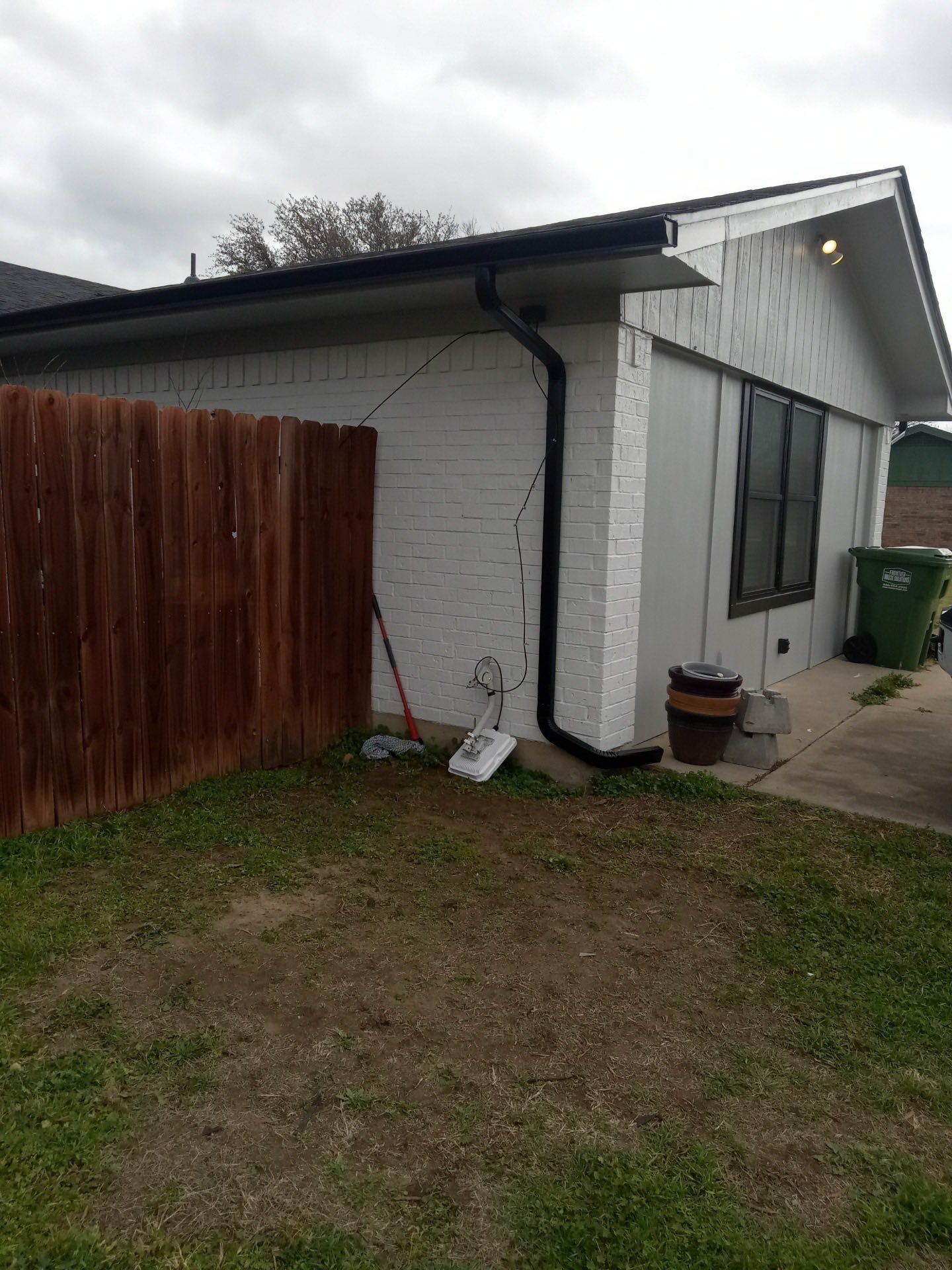 Backyard side view of a white house with black trim, wooden fence, grass, and a driveway with a green bin