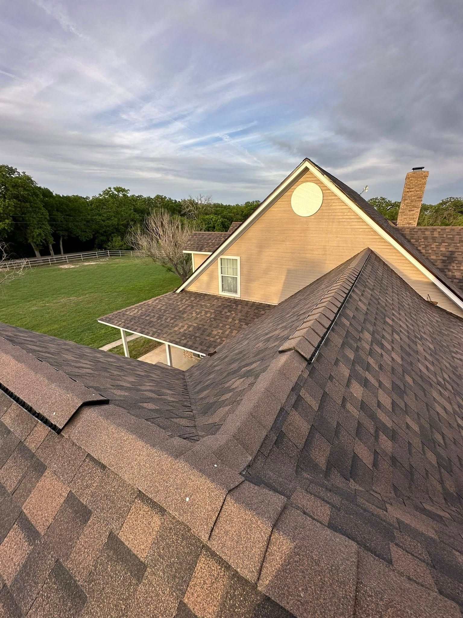 Roof shingles overlooking a tan house with a chimney and grassy yard under a cloudy sky