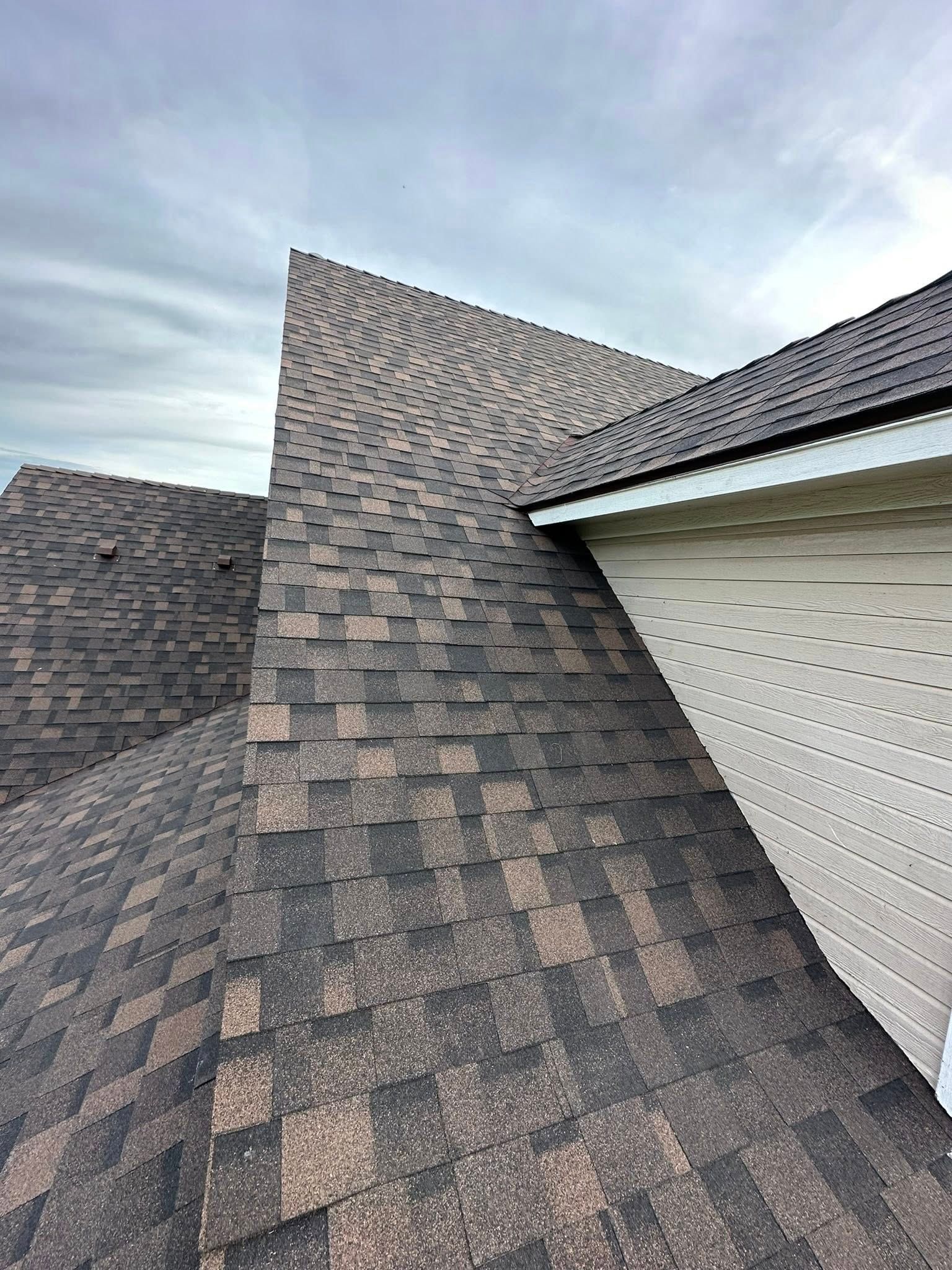 Dark gray shingled roof with a steep valley beside white siding under a cloudy sky