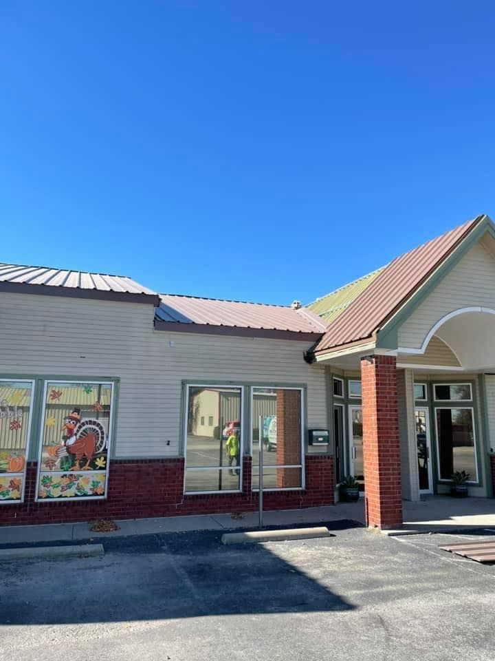 Strip-mall storefront with beige siding, brick columns, and colorful window posters under a clear blue sky