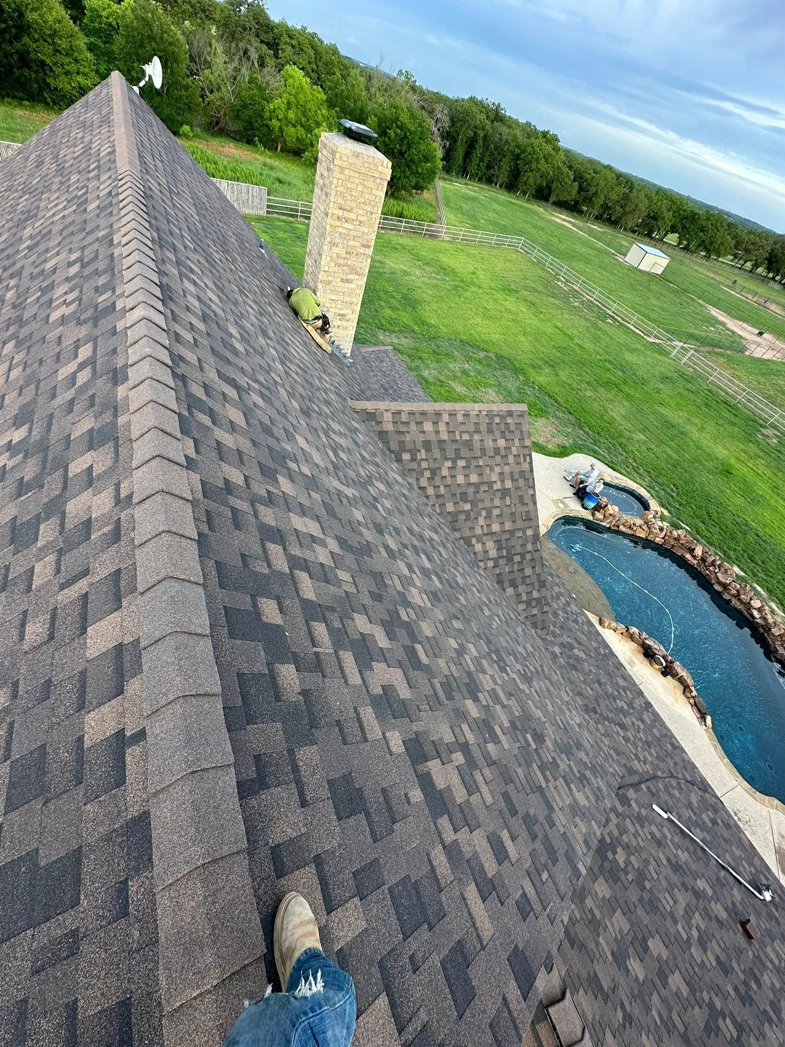 View from a shingled roof over a backyard with pool, stone chimney, and green lawn