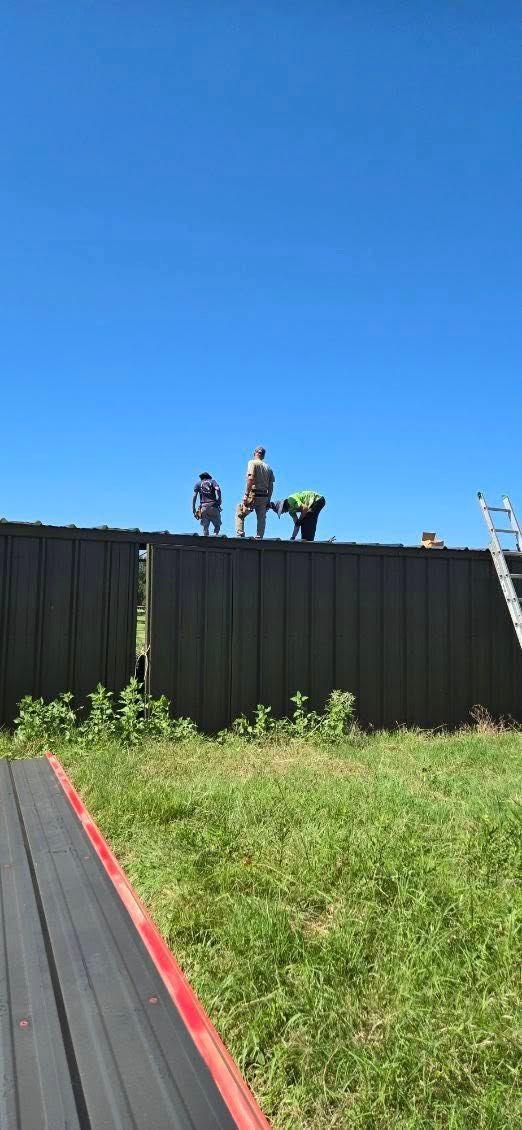 People working on a rooftop above a black fence, with green grass and blue sky in the foreground