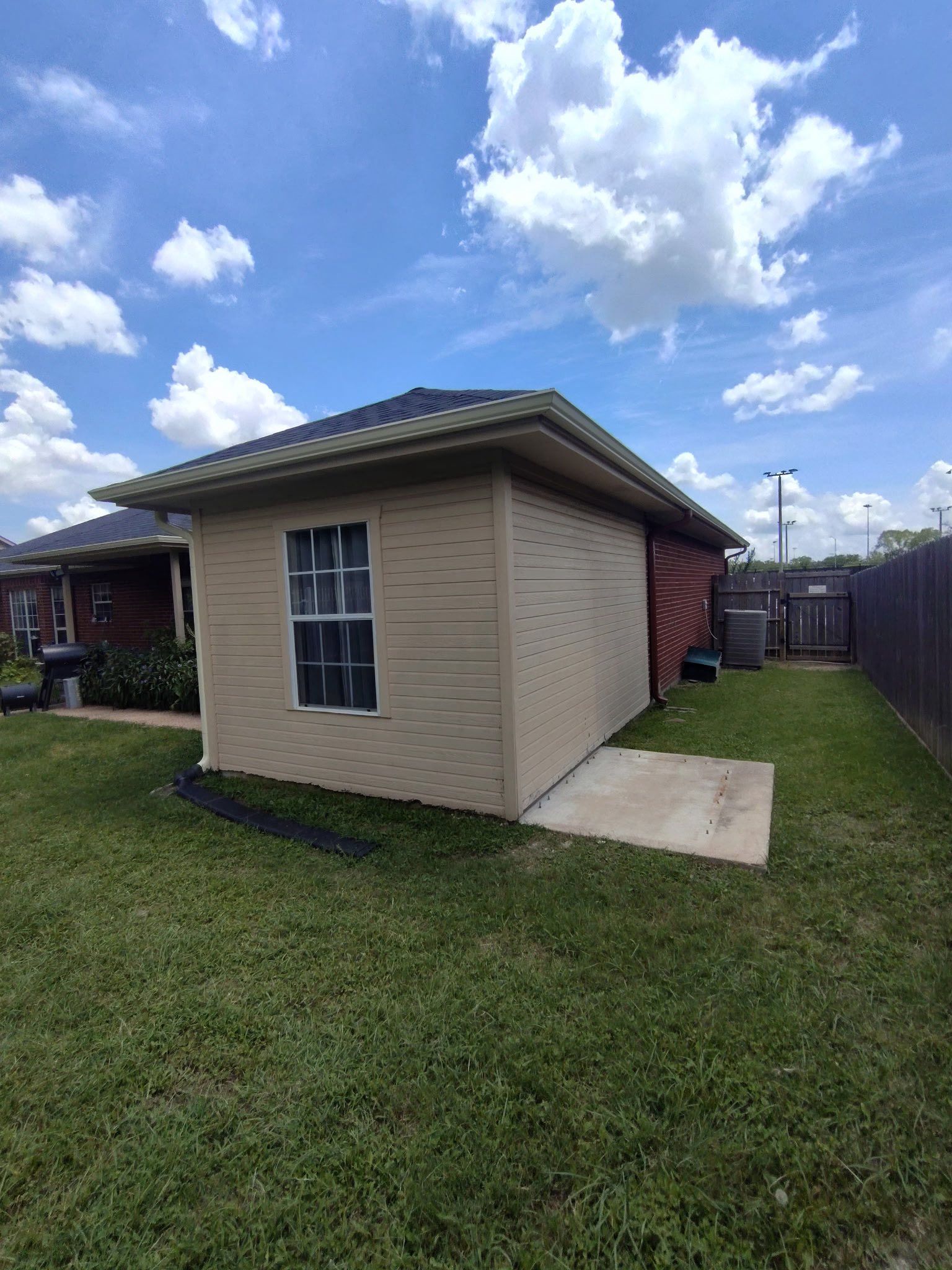 Small beige shed with a dark roof beside a concrete path in a grassy backyard under a blue sky.