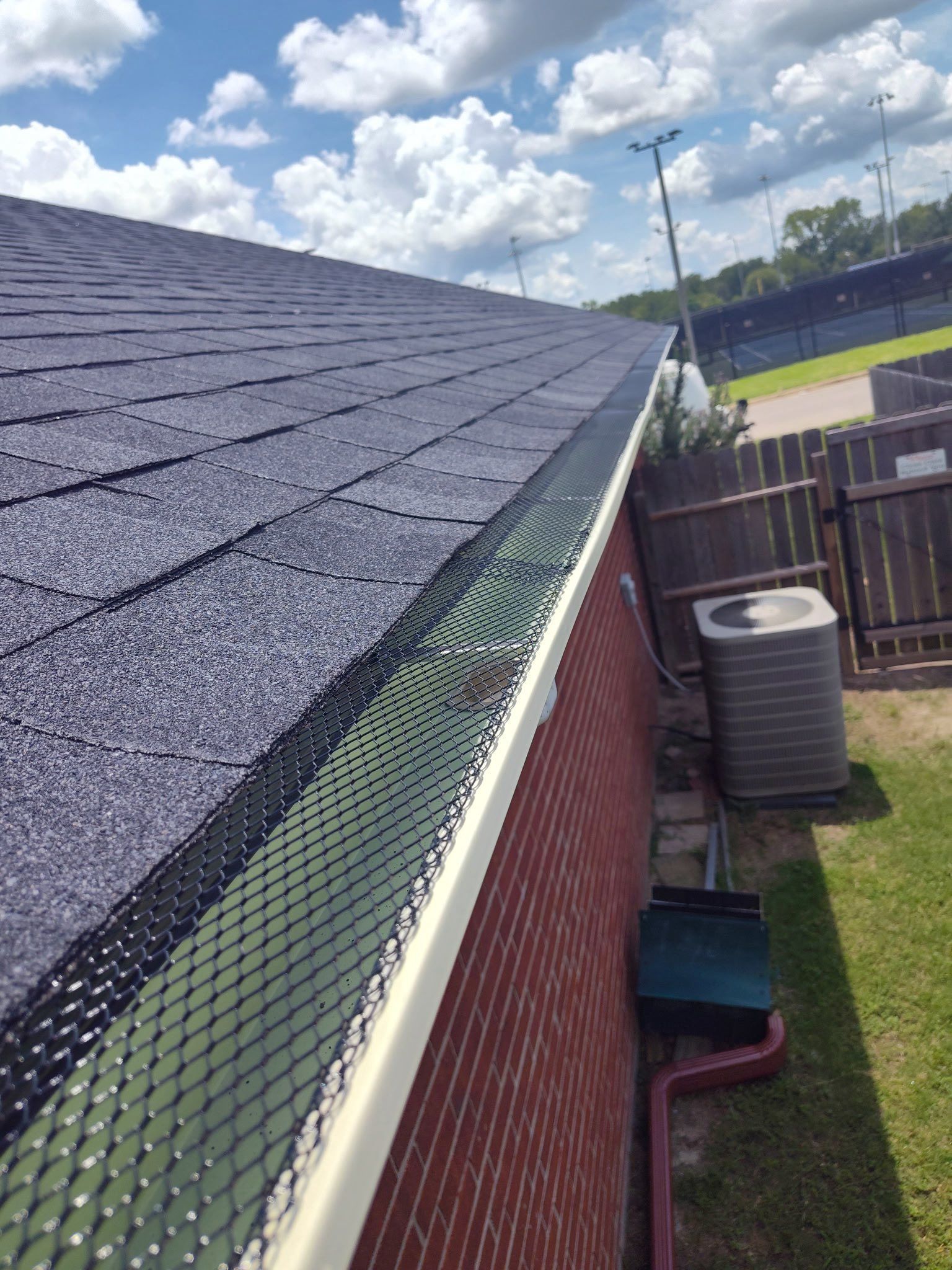 Roof with dark shingles, green gutter guard, red brick wall, and yard under a cloudy sky.