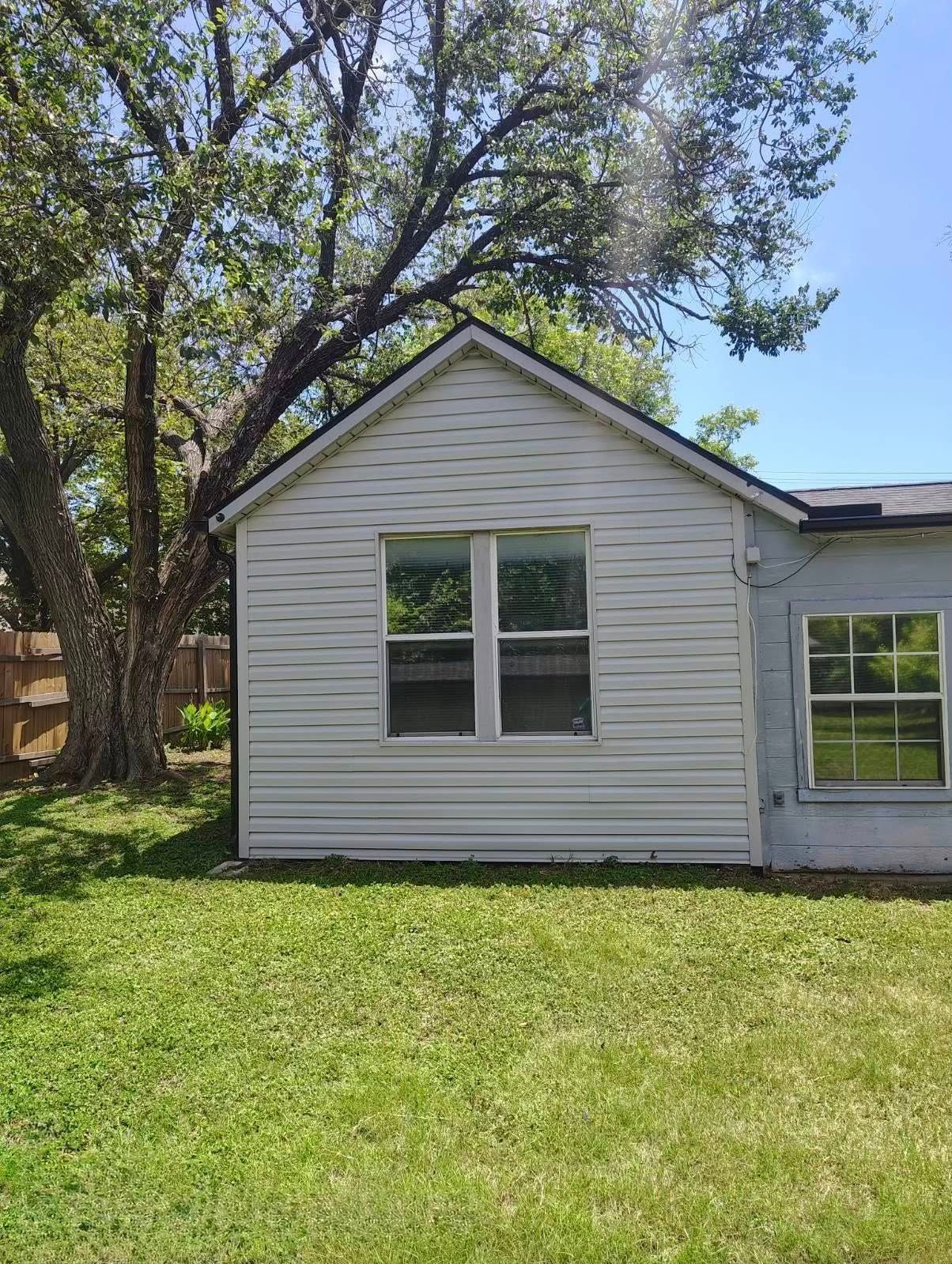 Small white house exterior with gabled roof, windows, and a grassy yard under trees