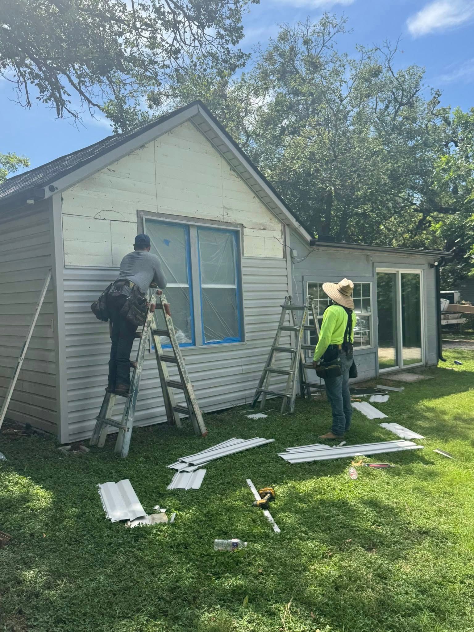 Two workers on ladders repairing the siding of a small white house on a sunny day.