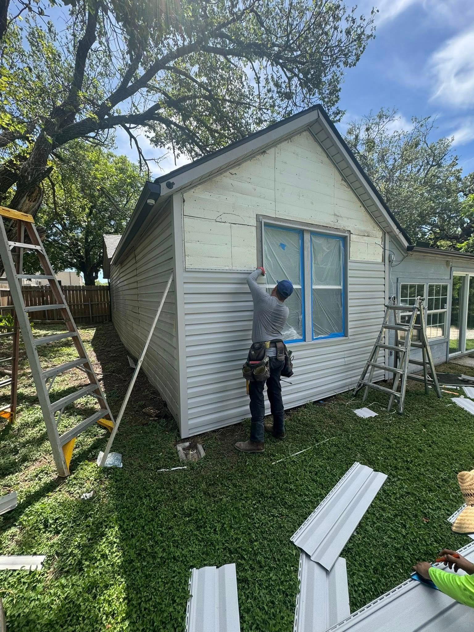 Person painting the trim of a small white shed with blue windows in a backyard.