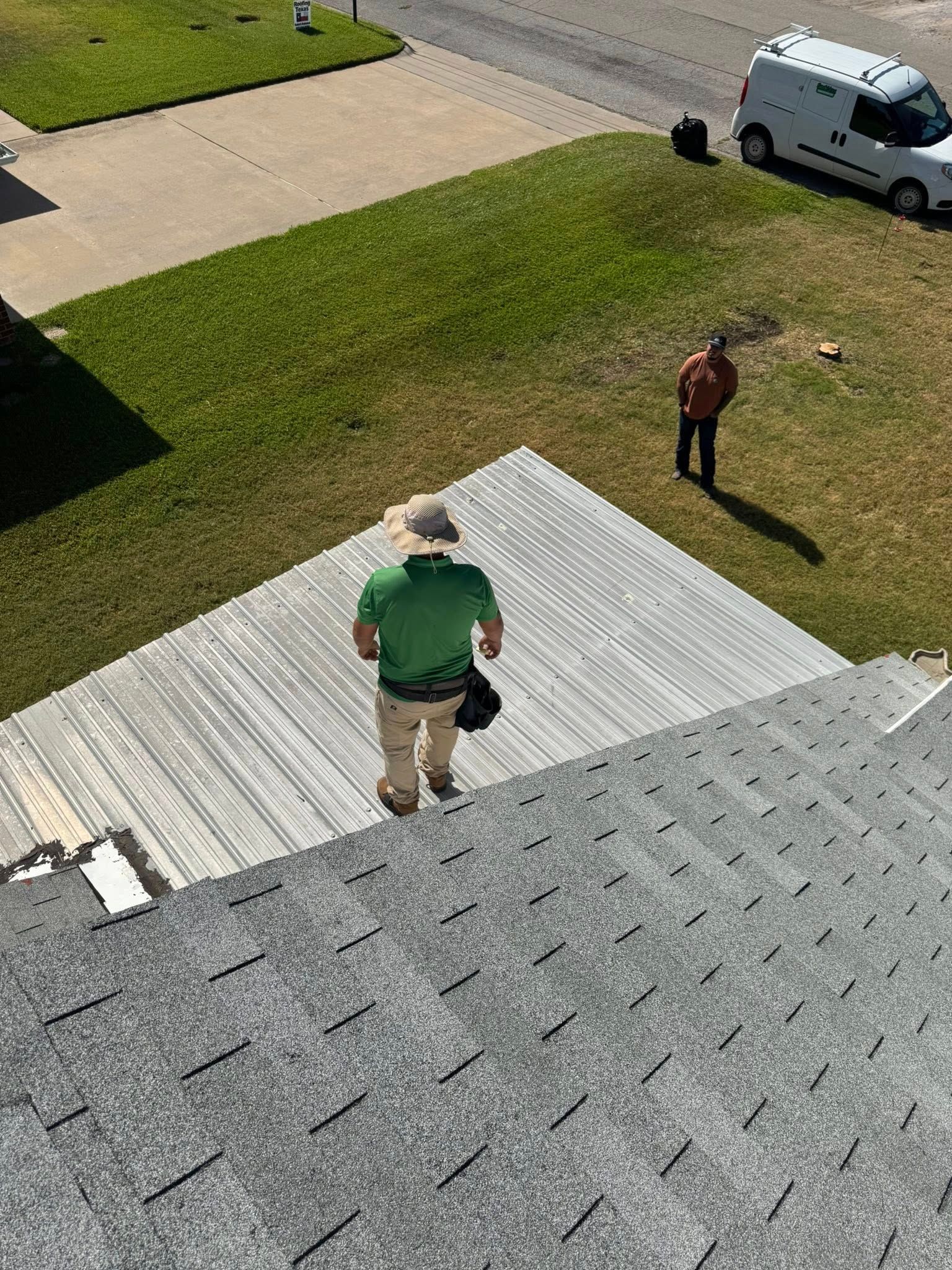 Two people standing on a rooftop near a driveway and van, with shingles and a ladder visible.
