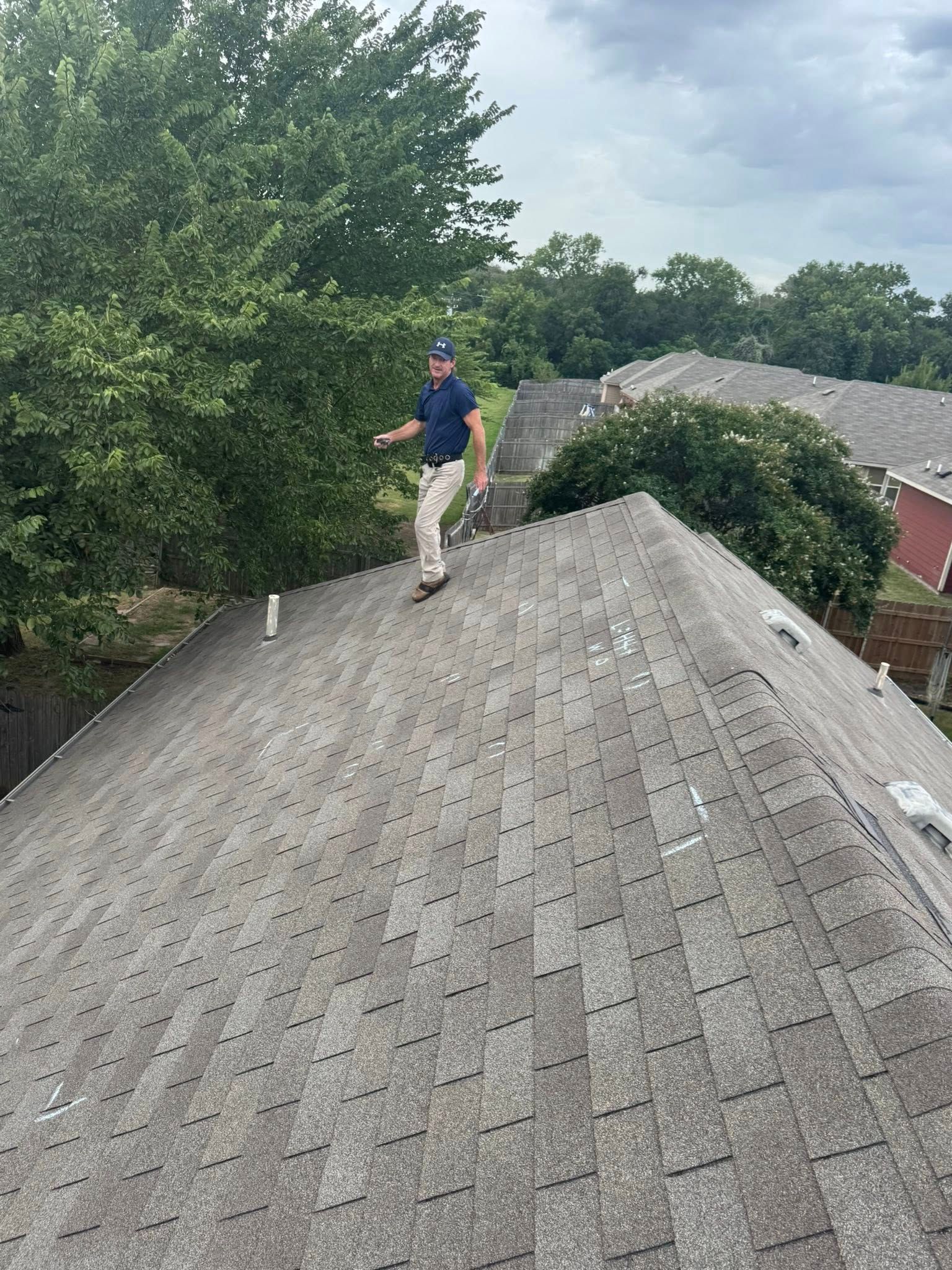 Person standing on a shingled roof beside trees and neighboring houses