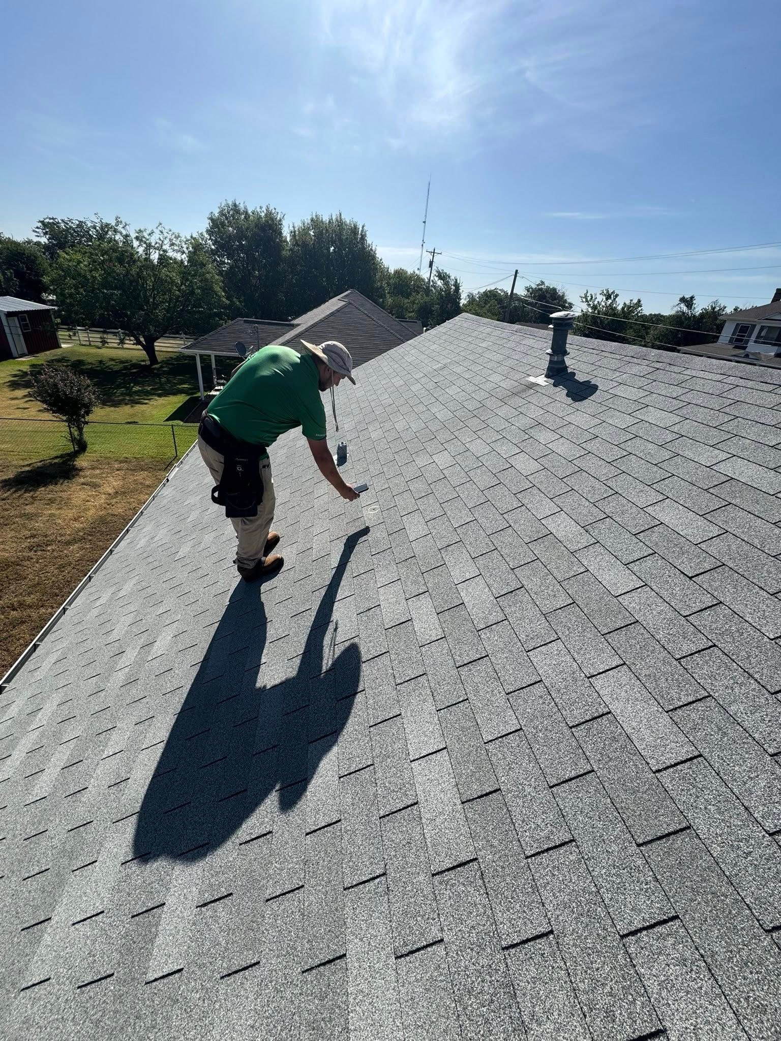 Worker in green shirt inspecting a shingled roof under bright sunlight.