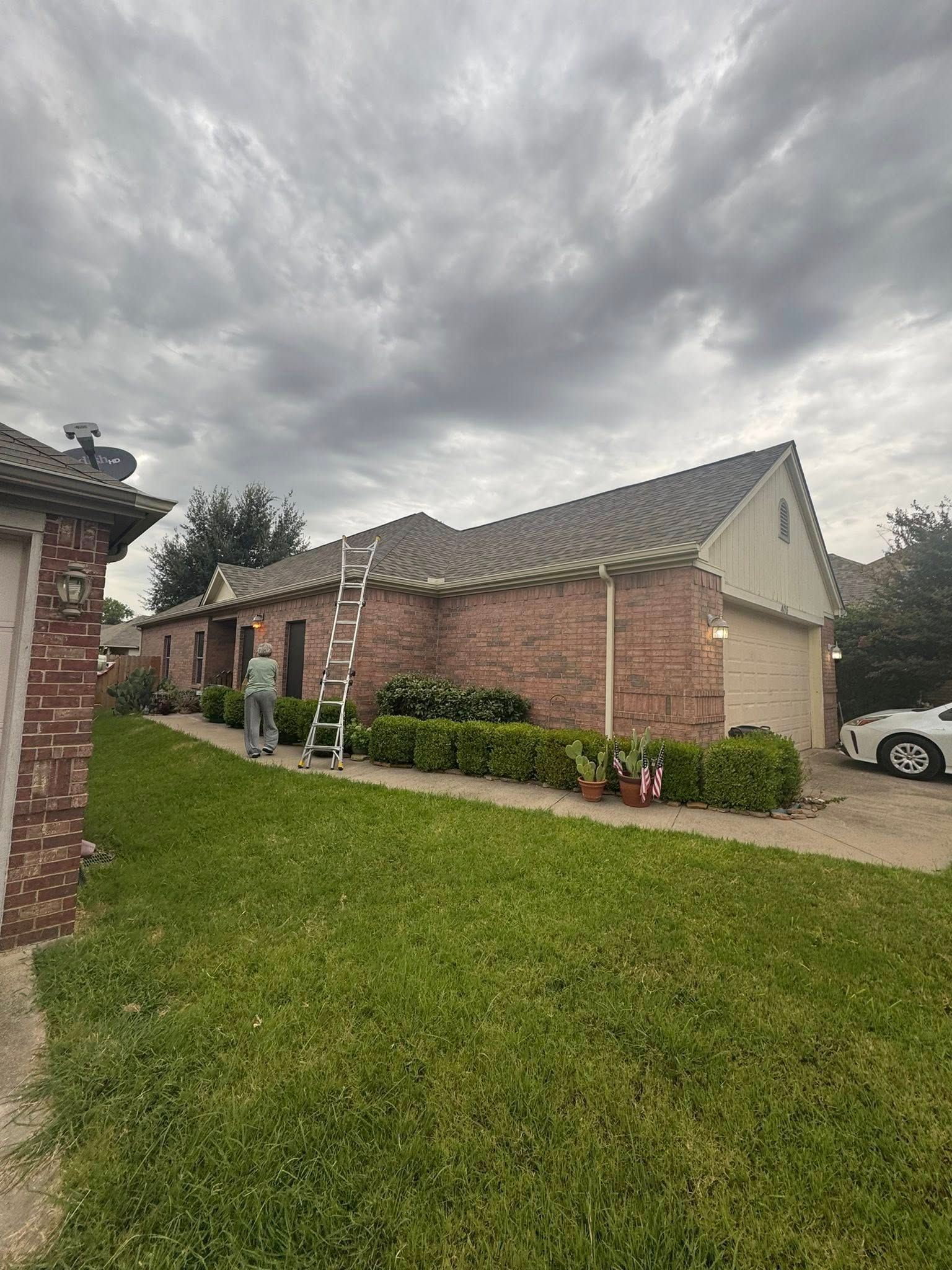 Brick house with gray roof and ladder in a grassy residential yard under cloudy skies