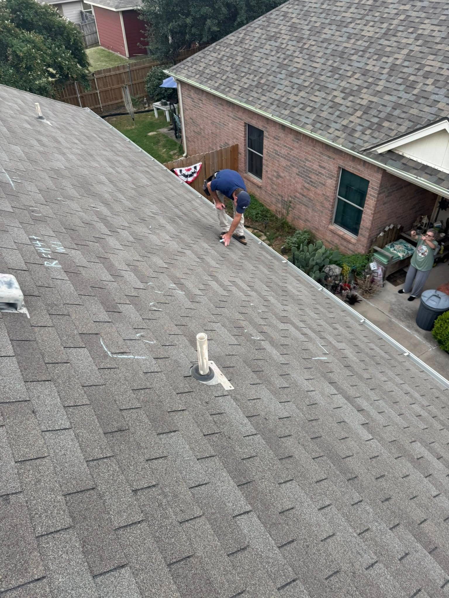 Worker repairing shingles on a house roof beside a brick home and outdoor walkway.